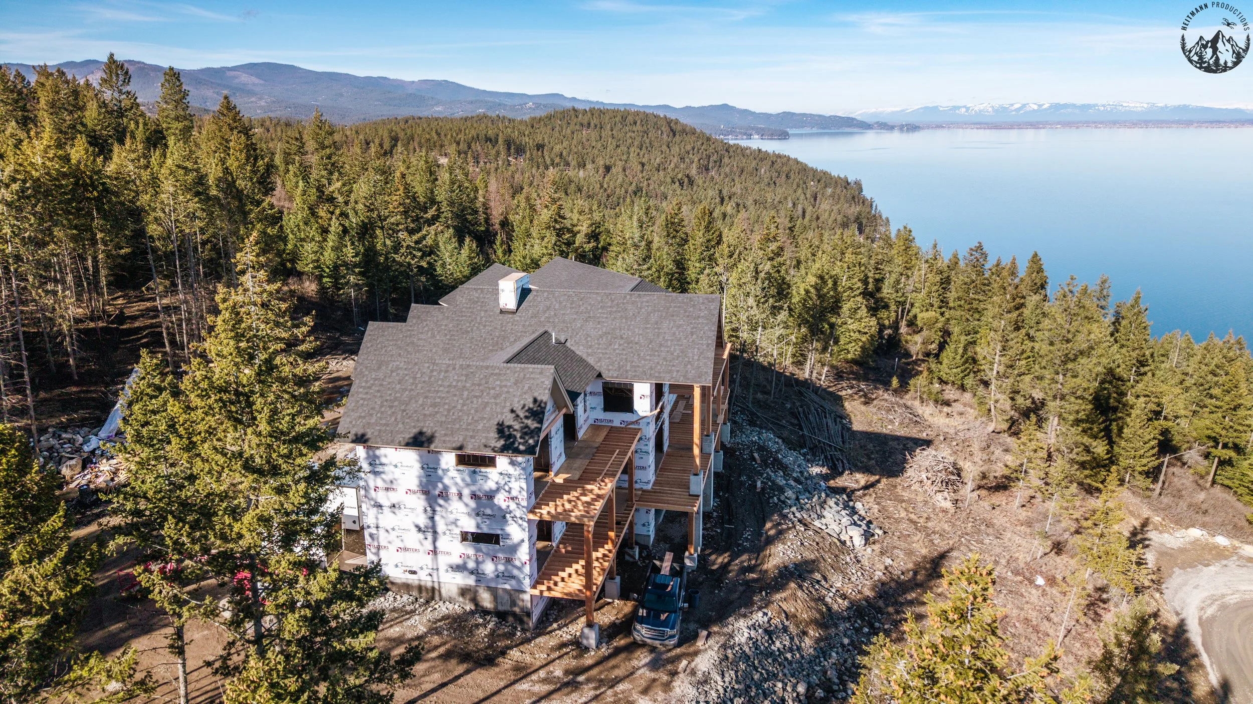 A house under construction on a hillside overlooking a large body of water and forested mountains.