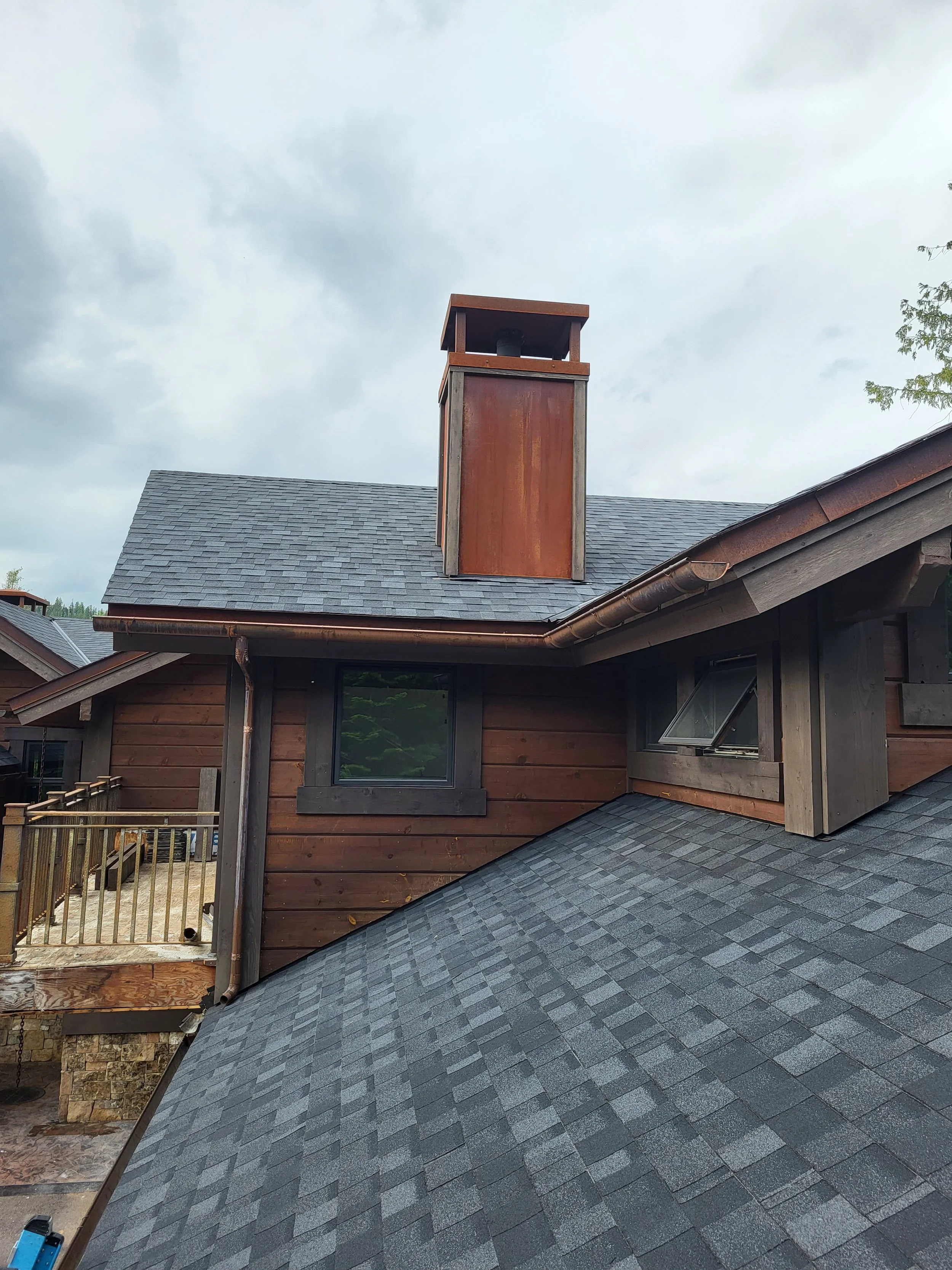 Close-up of a house rooftop with asphalt shingles and a rusty metal chimney on a cloudy sky day.