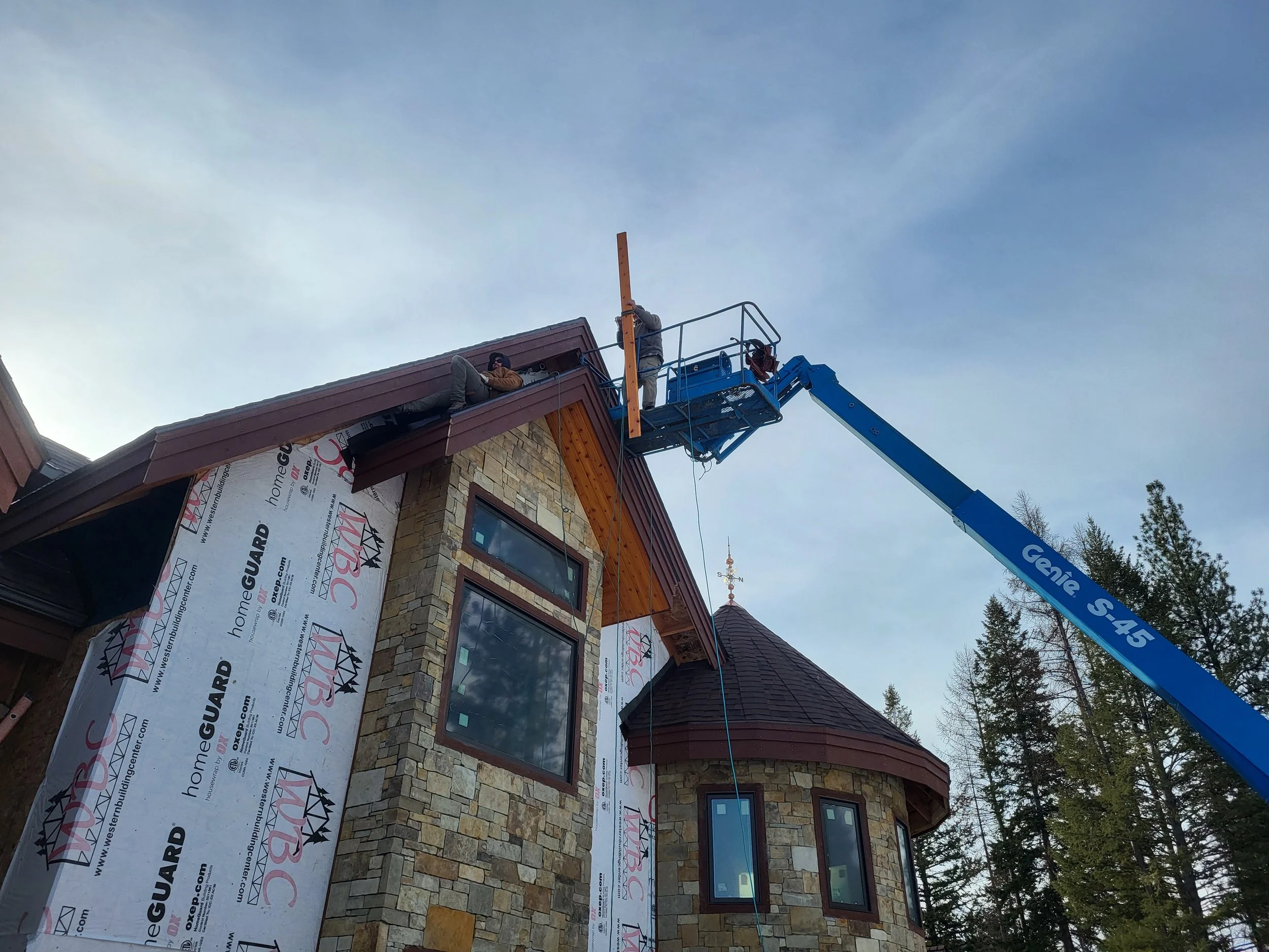 Construction workers are working on the roof of a stone house using an aerial lift. The house has a turret and large windows, with building wrap visible on part of the exterior. Trees are in the background under a partly cloudy sky.