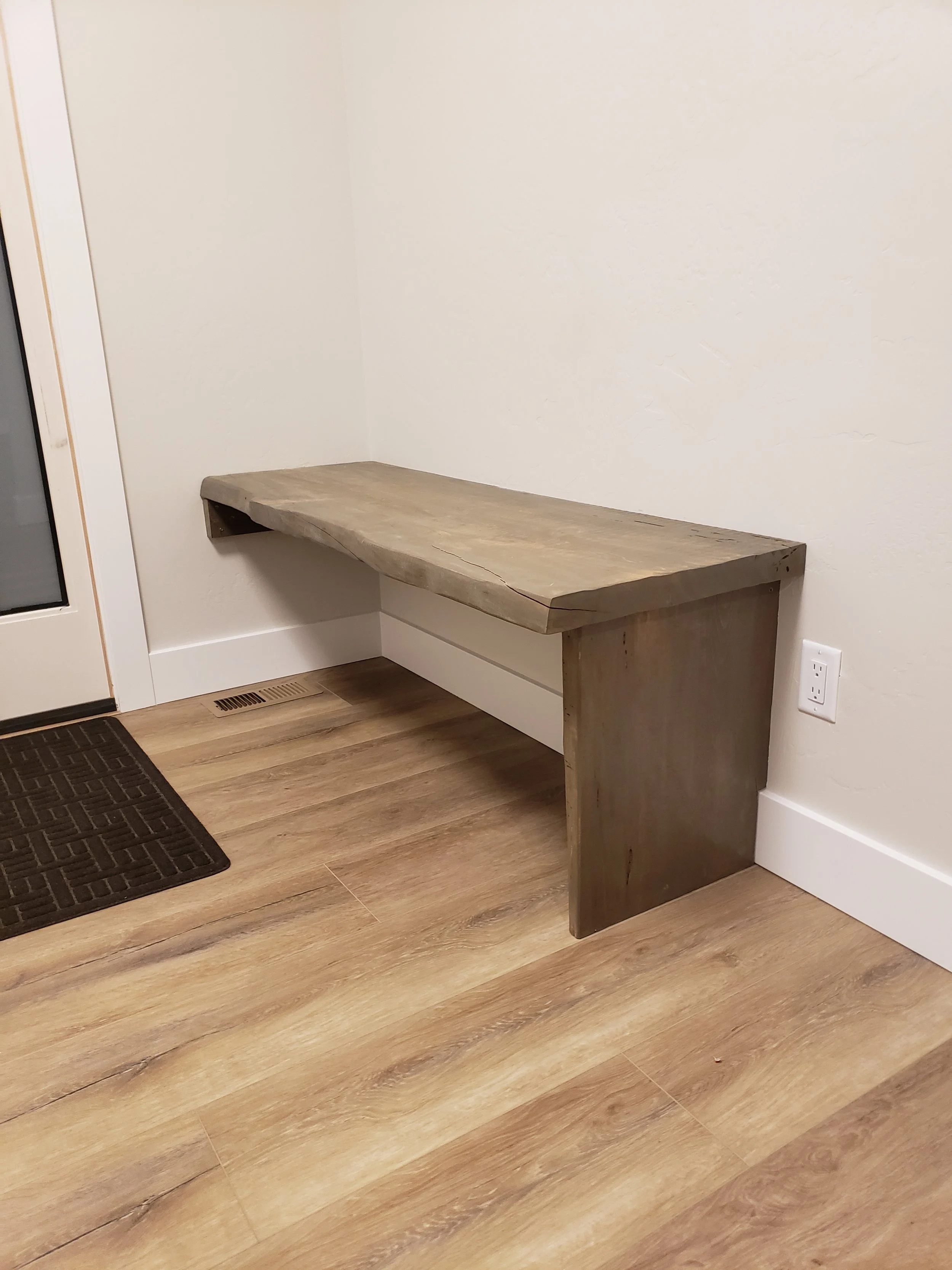 A rustic wooden bench with a thick, natural wood top and a single side support, situated in a corner of a room with hardwood flooring and a white wall.