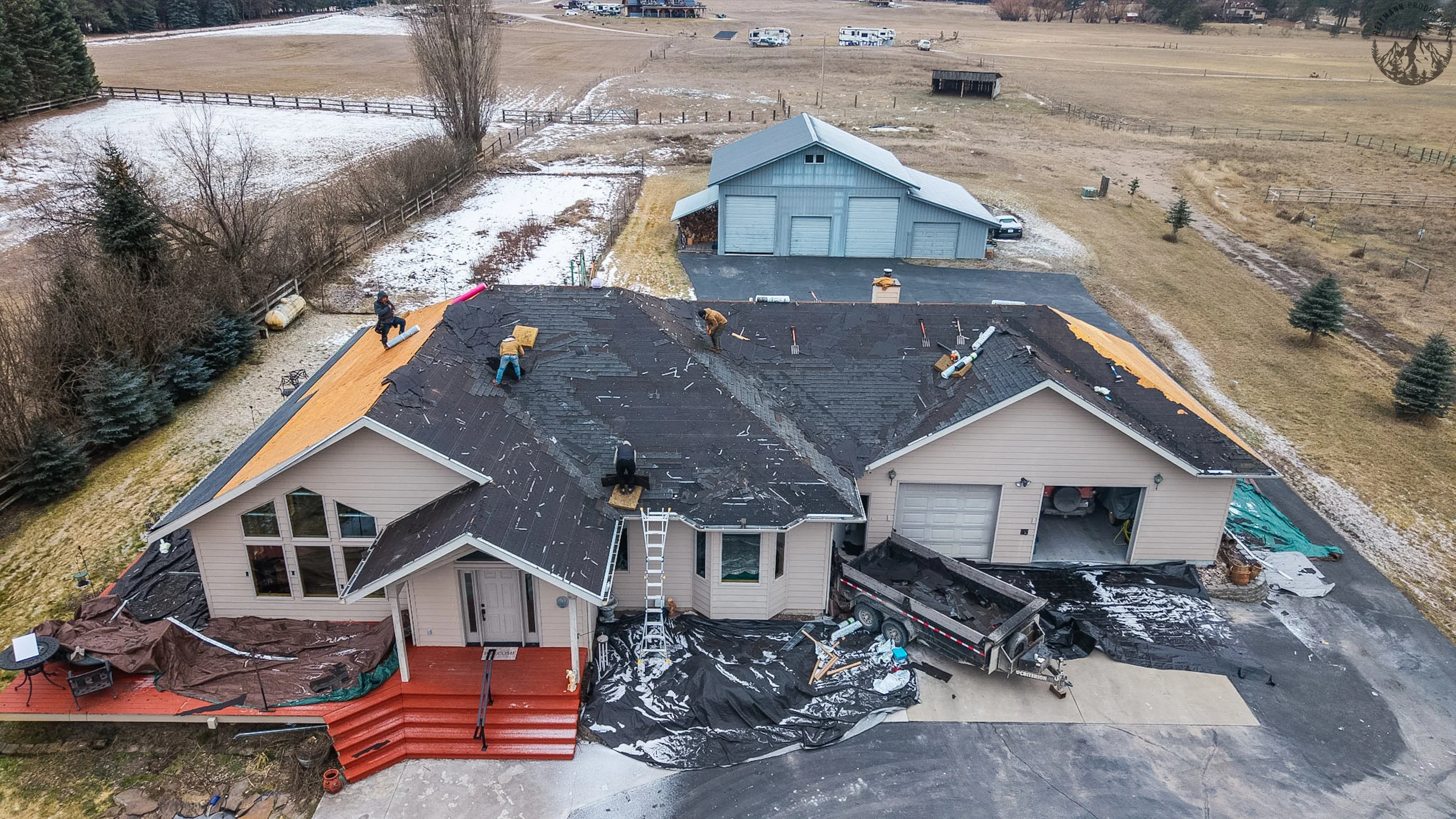 Aerial view of a house undergoing roof replacement, with workers removing old shingles and installing new roofing. The house has a beige exterior, a red front porch, and a driveway with a trailer. The backyard has some snow, a blue shed, and a fenced