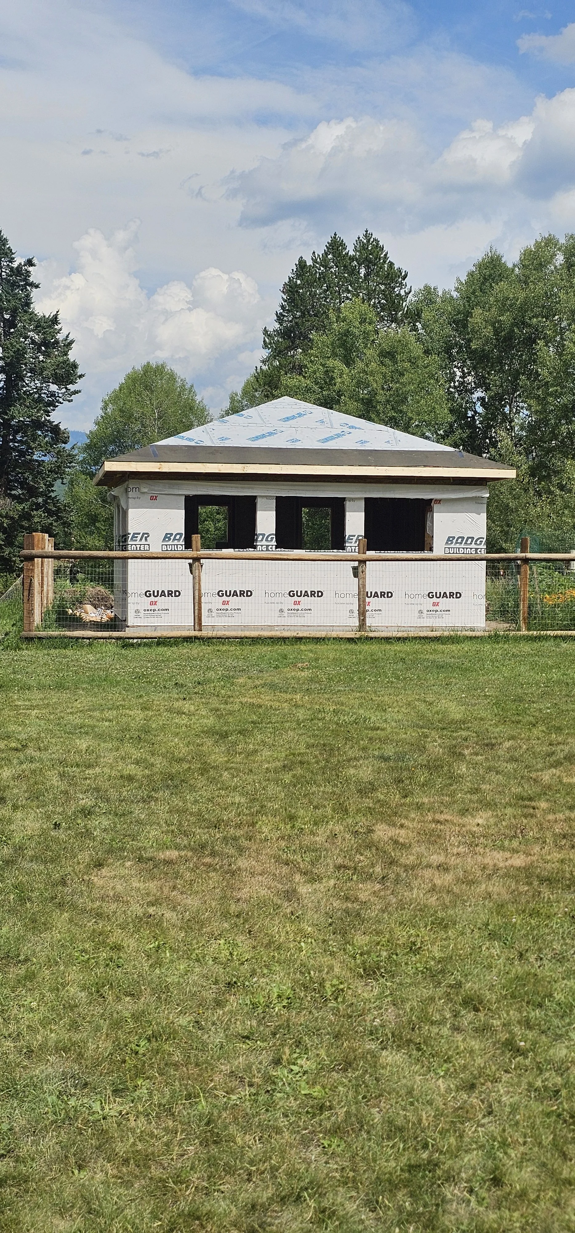 Construction site with a small building under construction, surrounded by a wooden fence, with trees and a partly cloudy sky in the background.