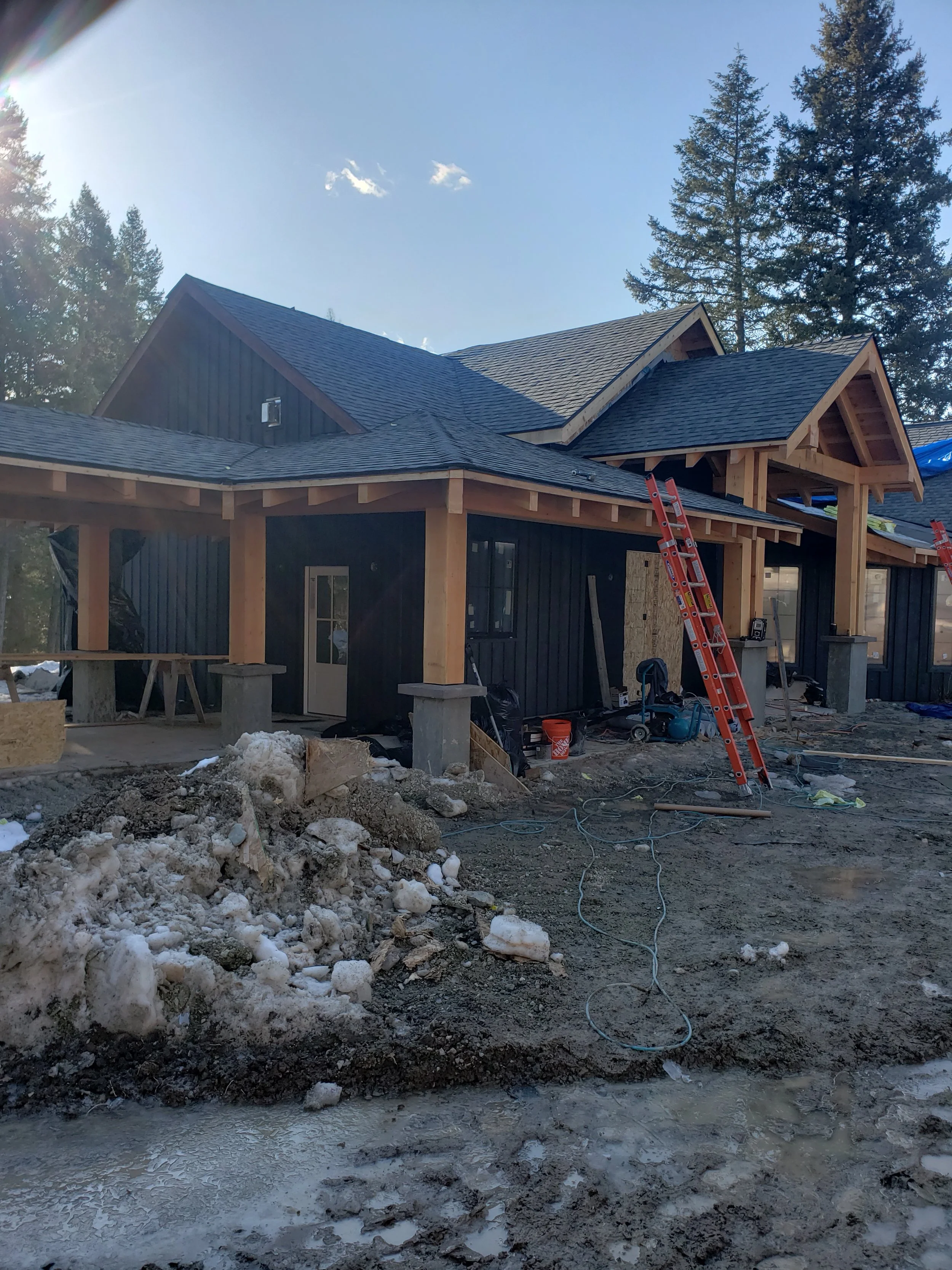 Construction site of a house with black siding, wooden beams, and roofing in progress; construction materials, tools, ladders, and debris are visible, with tall trees in the background.