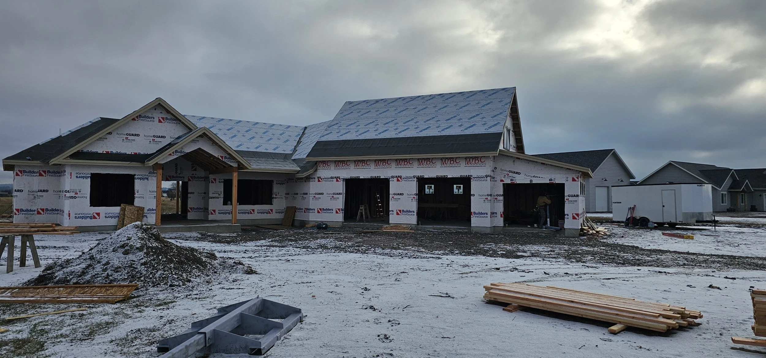 Under construction house with a snow-covered yard, partially finished siding, and roof framing, with nearby houses and construction materials on the ground.