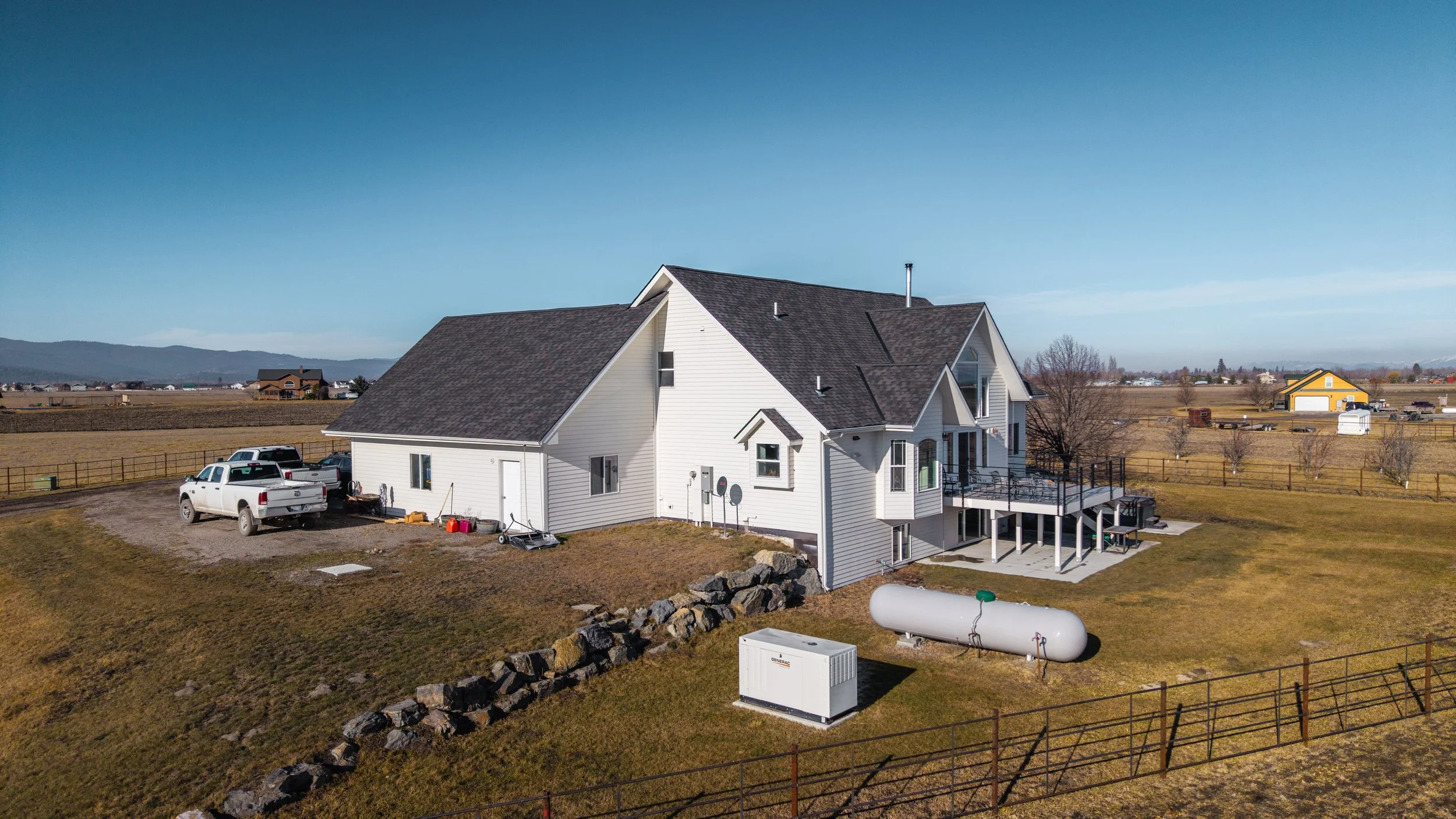 A large white house on a rural property with a brown roof, surrounded by an open yard and fenced fields. Several vehicles are parked near the house, and there are outdoor equipment and a propane tank visible. The landscape features distant hills and 