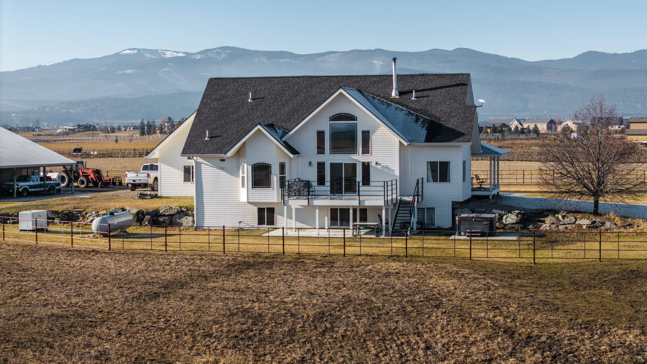 A large white house with a black roof, situated on a grassy, fenced property with a mountain range in the background.