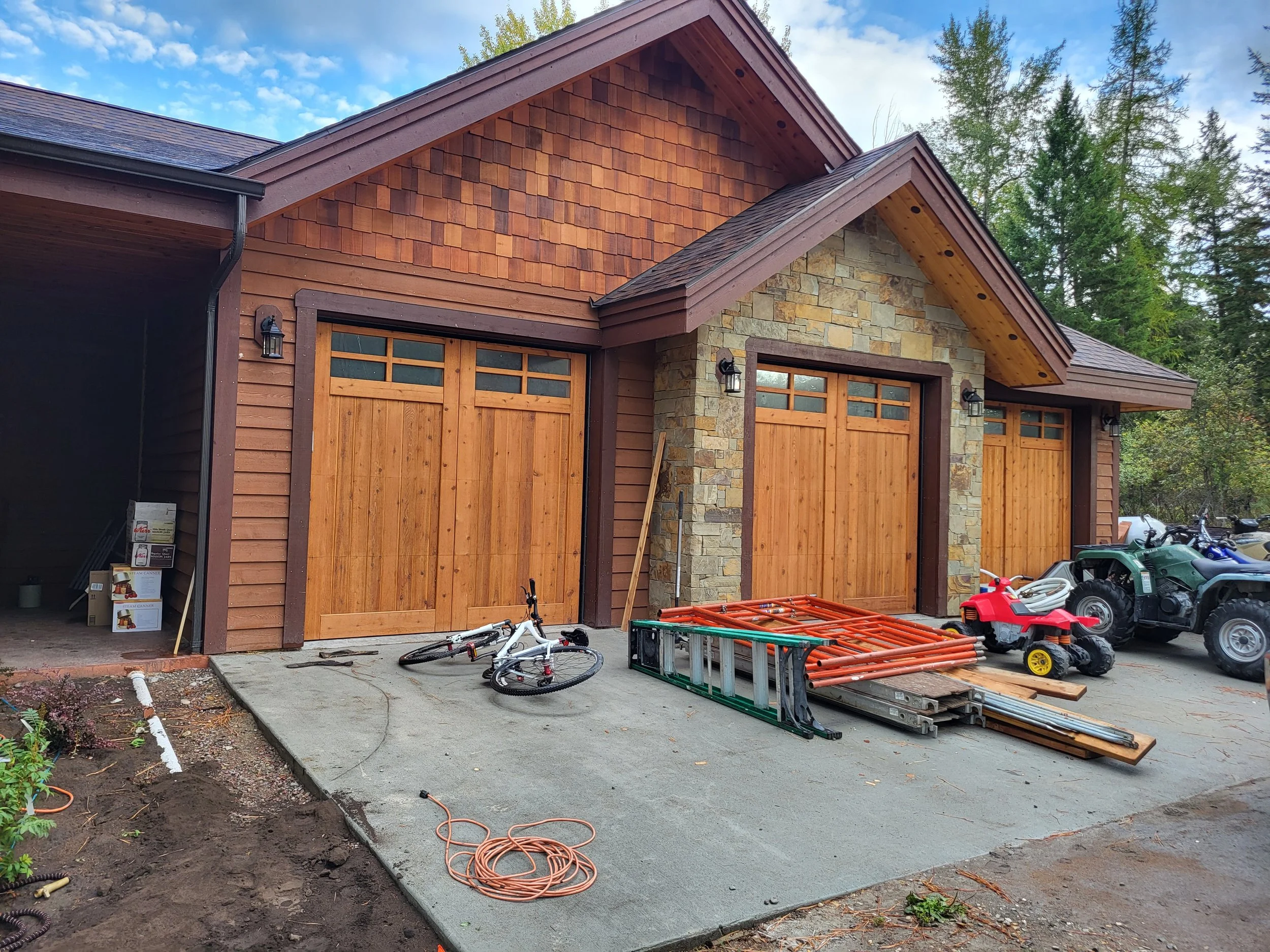 Three wooden garage doors in front of a house with stone and wood exterior; construction tools and materials are on the driveway, including a bicycle, ladders, and a toy vehicle.
