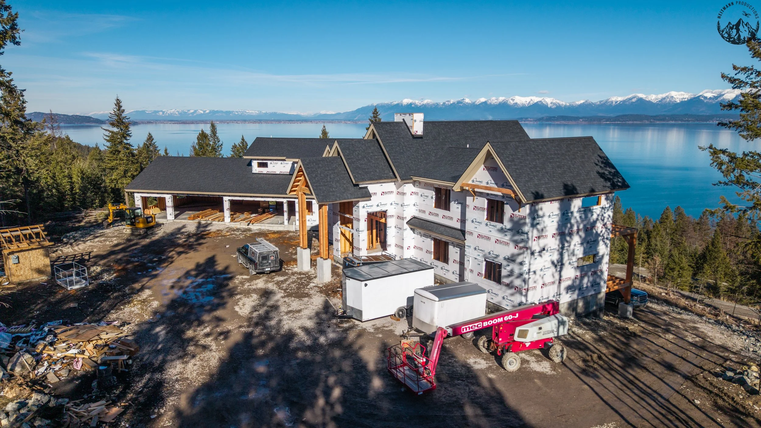 Construction site of a house with black roofing tiles in progress, overlooking a lake and snow-capped mountains, with construction equipment and materials around.