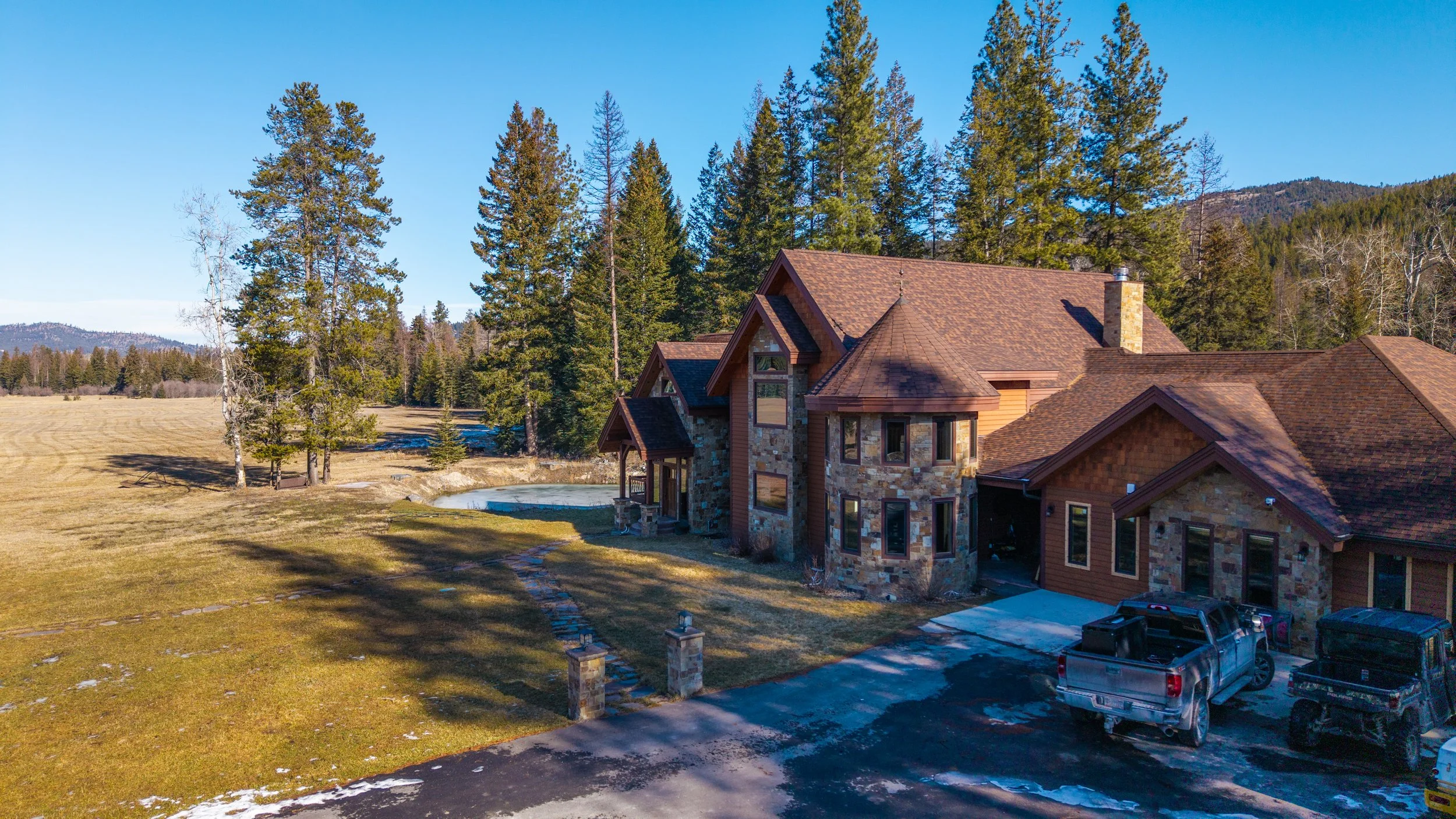 A large house made of stone and wood situated in a scenic area with tall pine trees and mountains in the background, with parked trucks in the driveway.