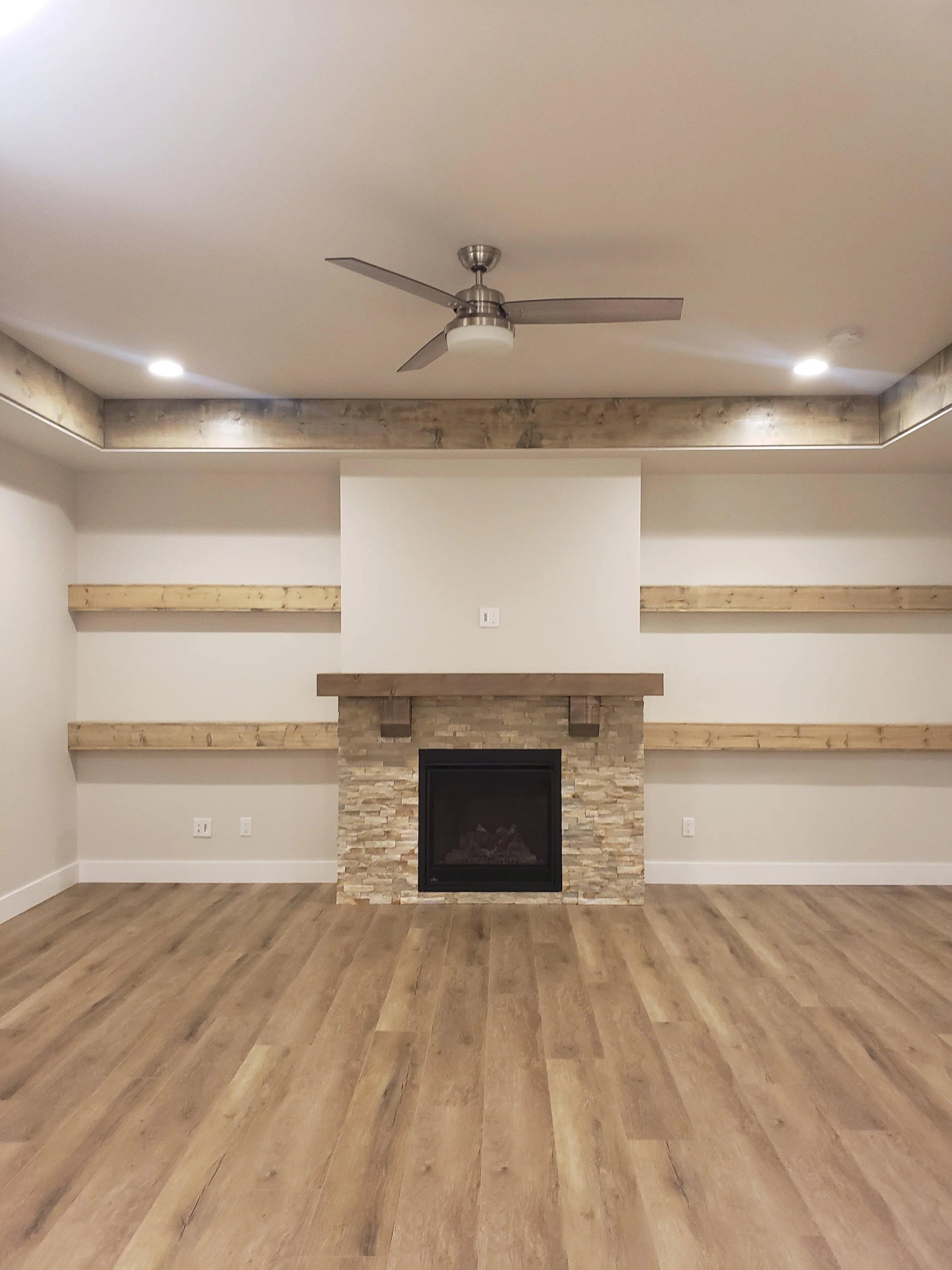 Empty living room with hardwood floors, a stone fireplace, held up by a wooden beam, and two wooden shelves on each side, with a ceiling fan and recessed lights.