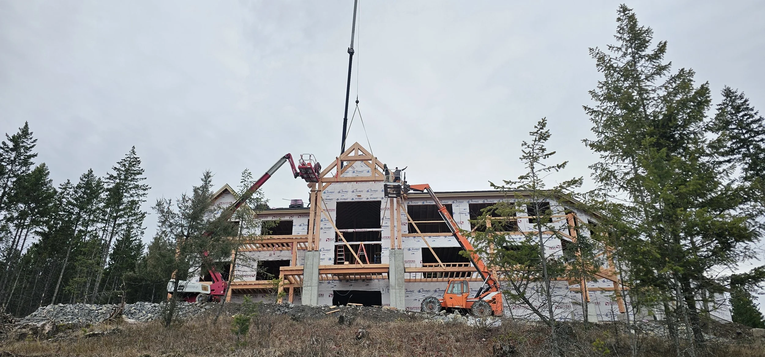 Under construction house with wooden framework on a hillside surrounded by trees, with construction cranes and equipment present.