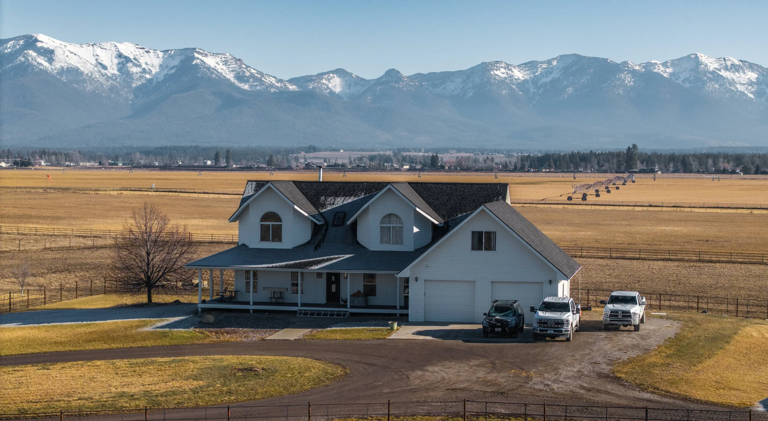 White two-story house with a front porch, surrounded by a lawn, with mountains in the background and a large field with hay bales and snow-capped peaks.