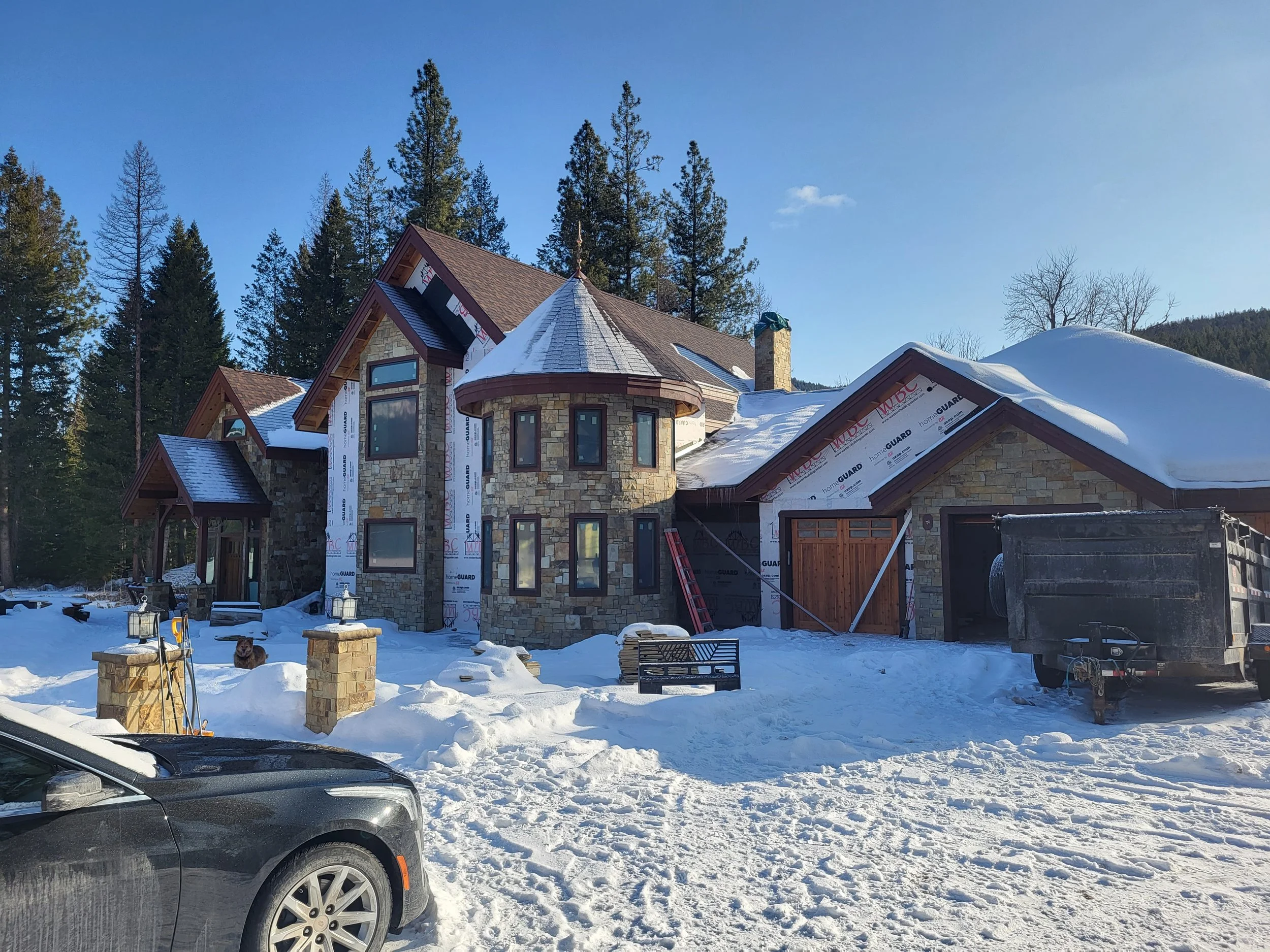 A large stone house under construction surrounded by snow, with tall pine trees in the background and a black vehicle in the foreground.