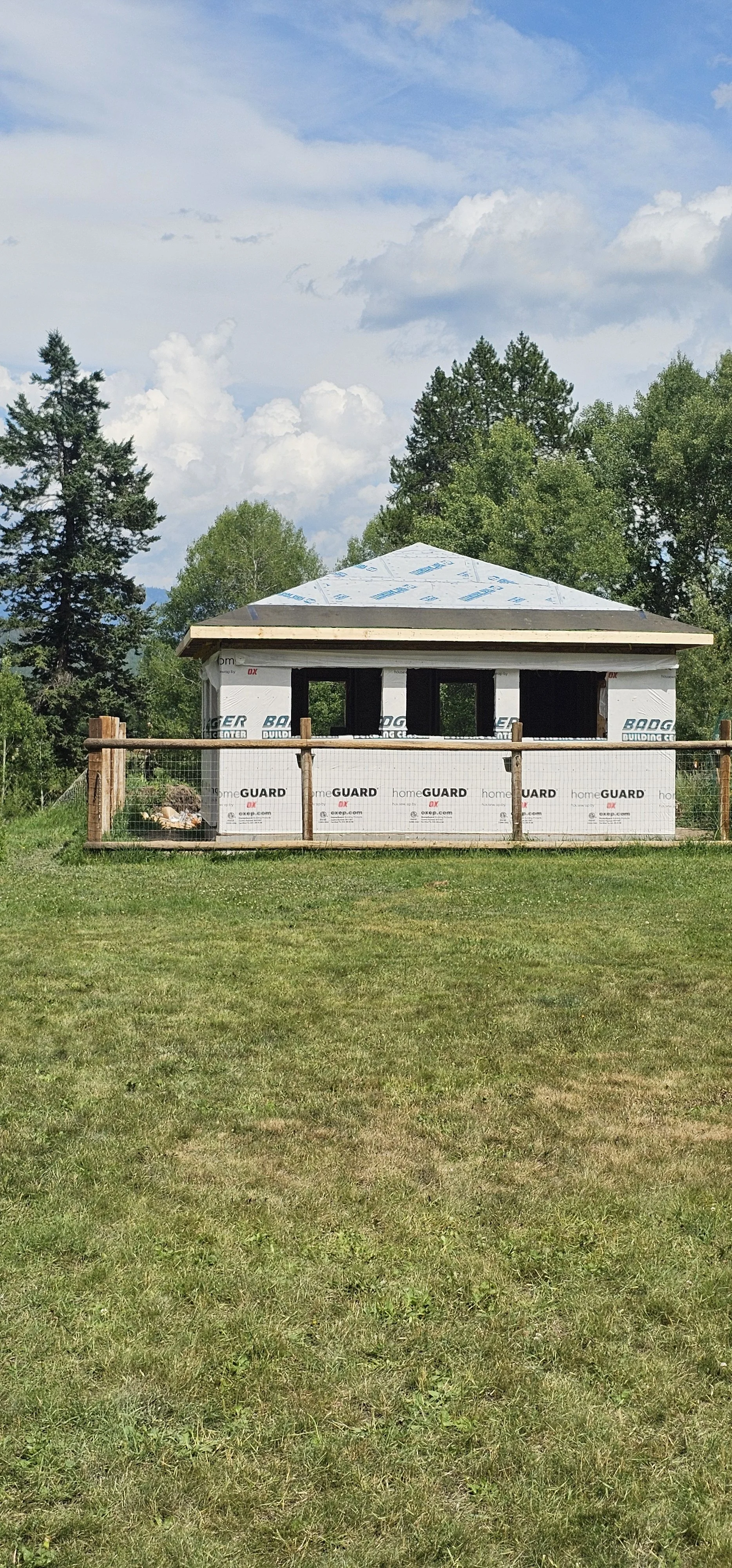 A house under construction with black window frames and a gray roof, surrounded by a wooden fence, in a grassy area with trees and clouds in the sky.