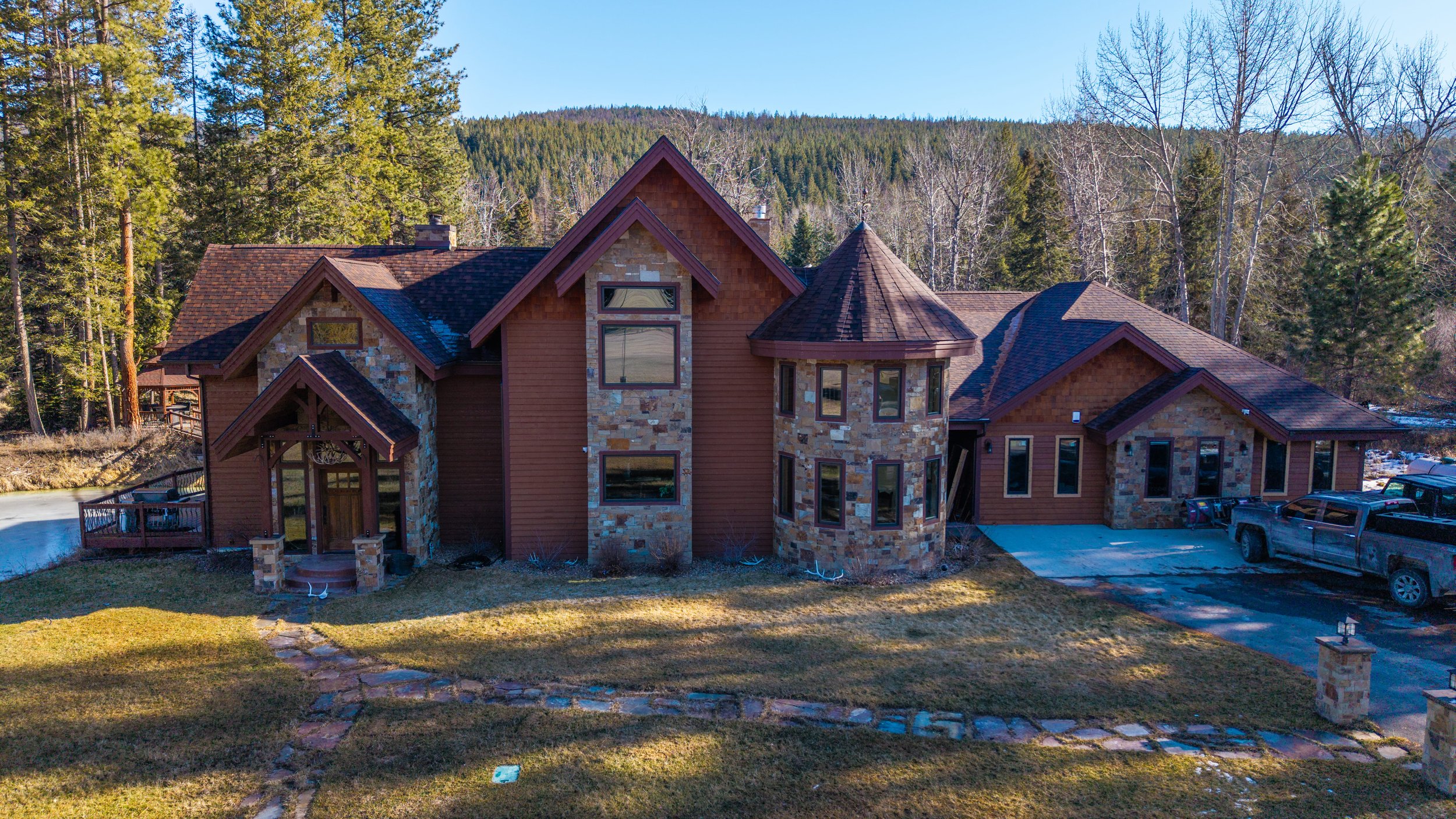 Large house with a stone and wood exterior, situated in a wooded area, with a driveway and parked trucks, and a stone pathway leading to the front door.