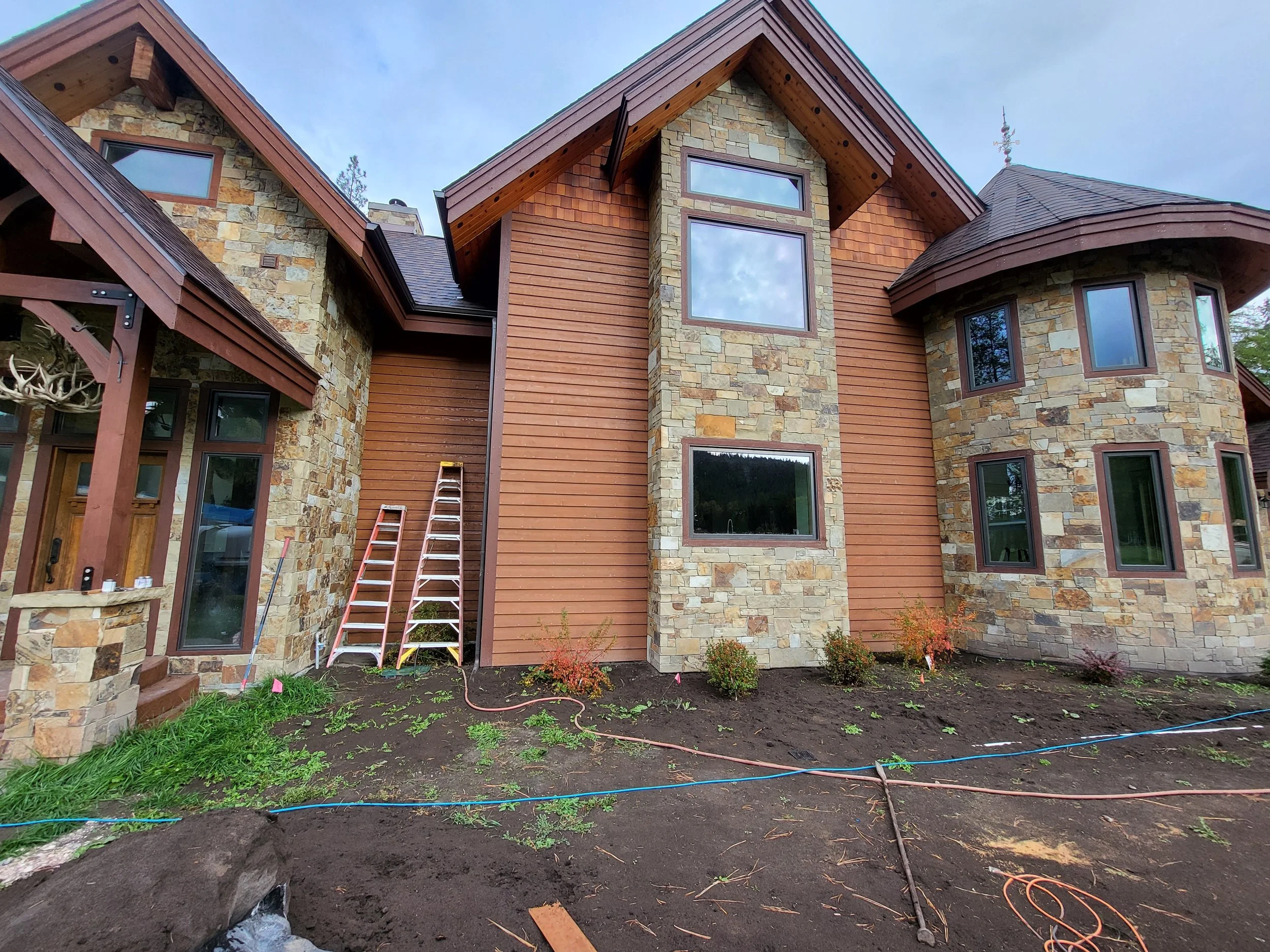Front view of a partially constructed house with stone and wood siding, multiple windows, and scaffolding, with dirt and plants in the yard.