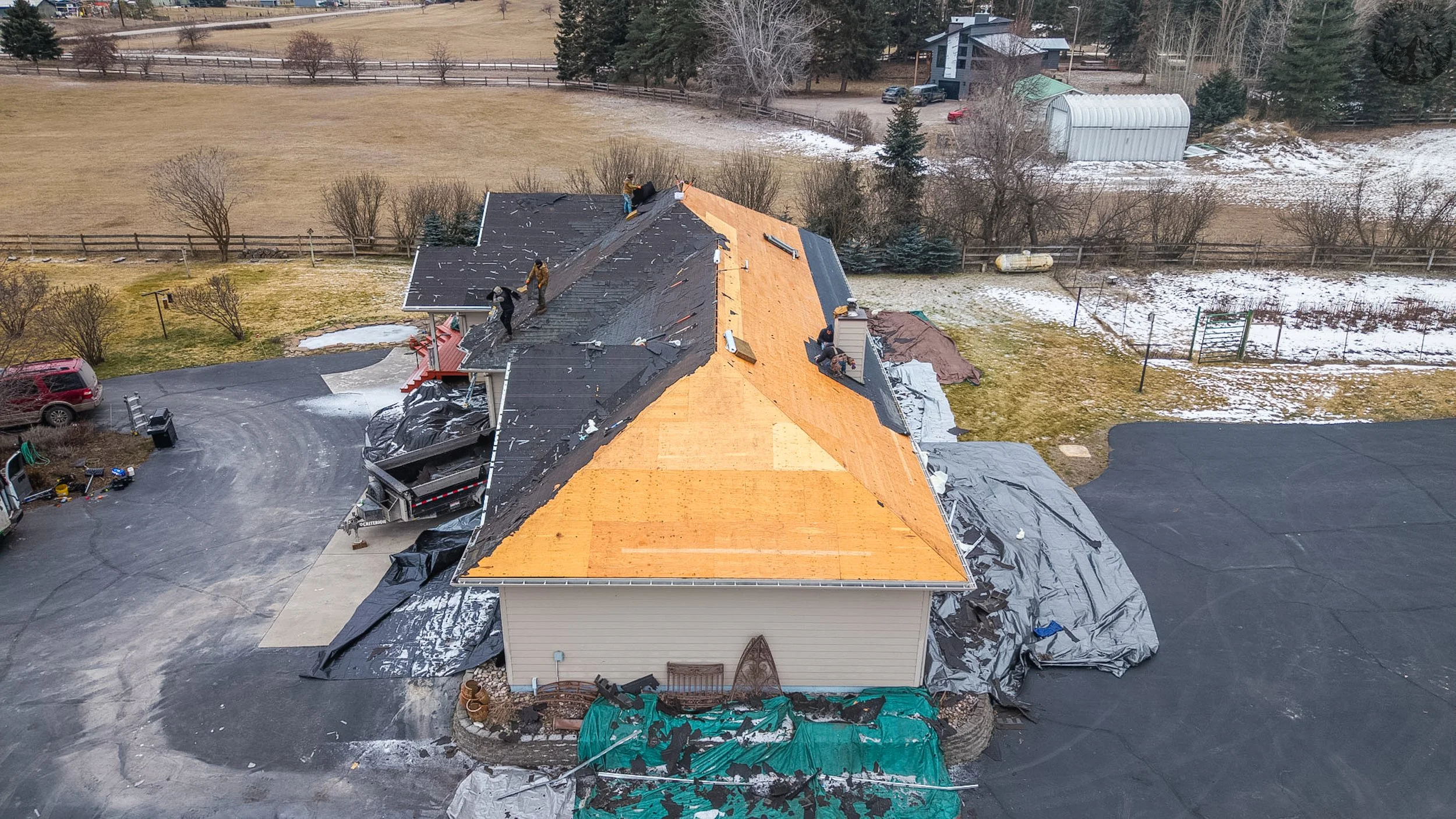 Aerial view of a house undergoing roofing renovation with workers replacing the shingles, some of which are already removed, exposing the plywood sheathing. The house is surrounded by a driveway, yard, and nearby snow patches.
