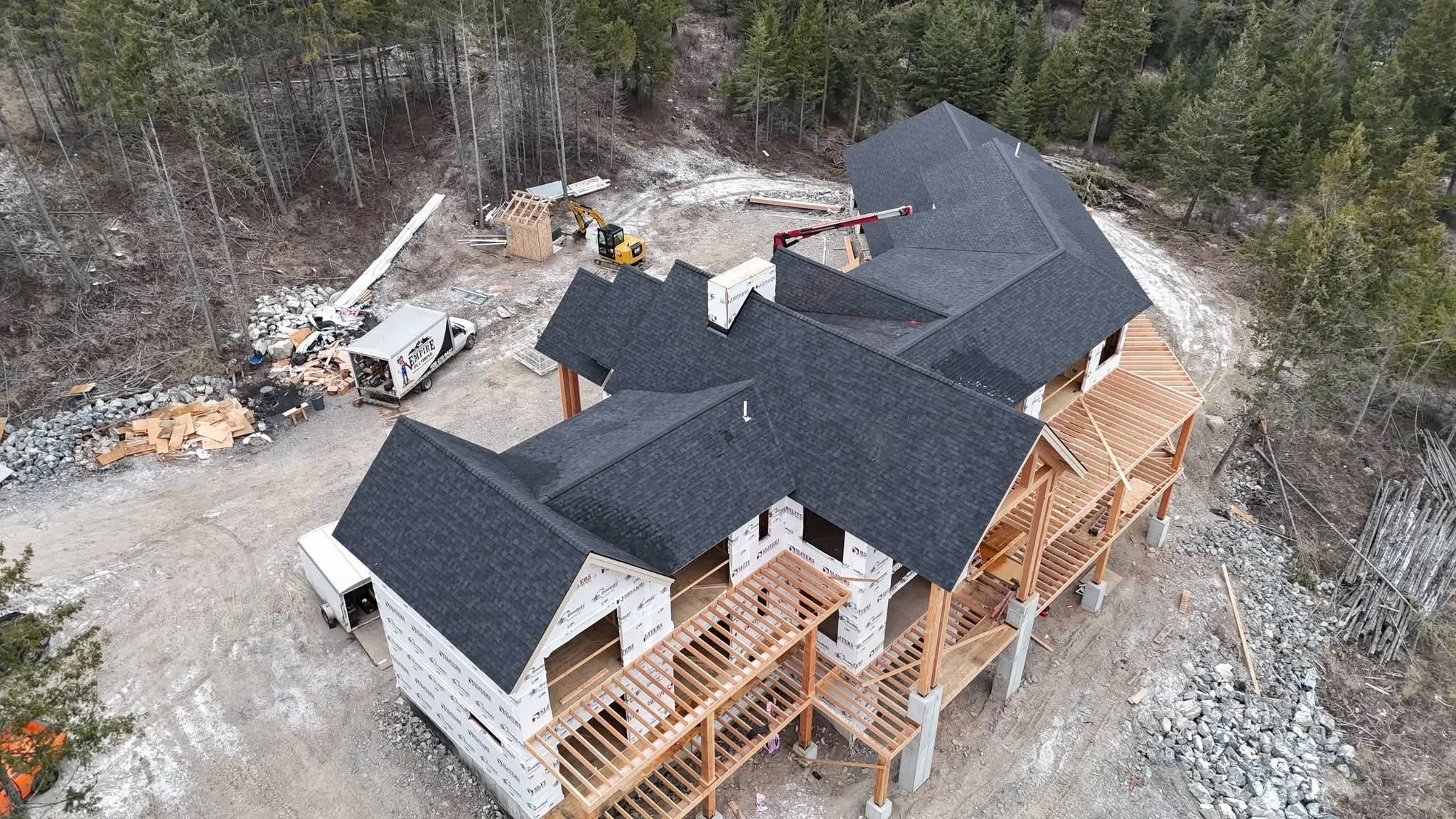 An aerial view of a house under construction in a wooded area. The house has black roofing, wooden framing, and some exterior walls wrapped in white. Construction equipment, a truck, and building materials are visible around the site.