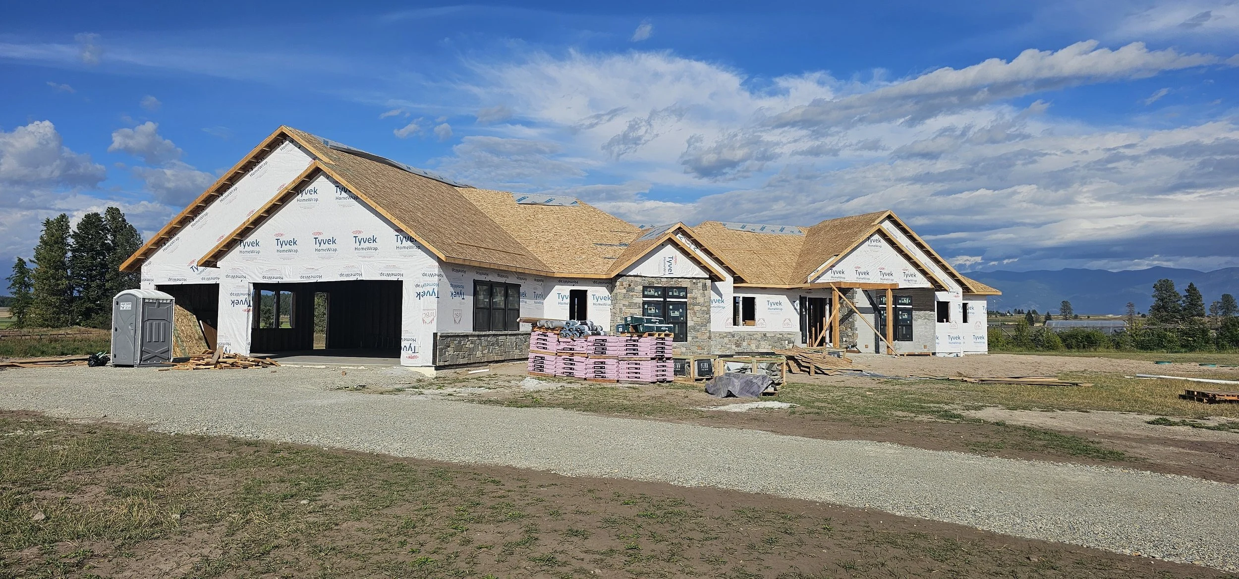 Under-construction house with a wooden roof, brick walls, and construction materials in front, on a partly cloudy day.