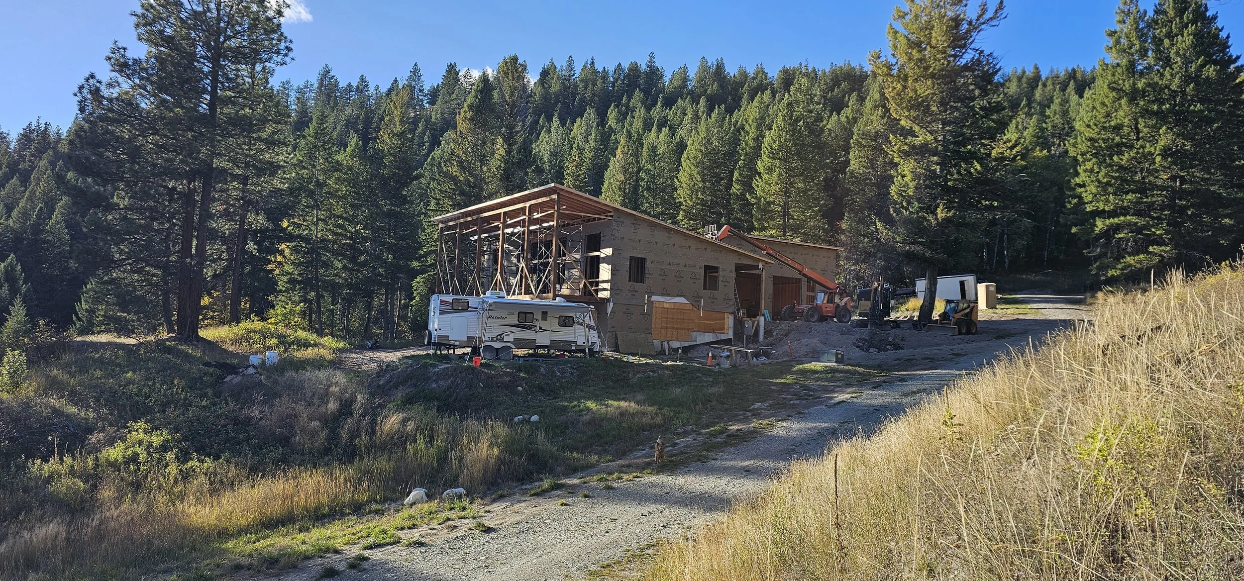 Under construction house in a wooded area with construction equipment and a trailer parked nearby.