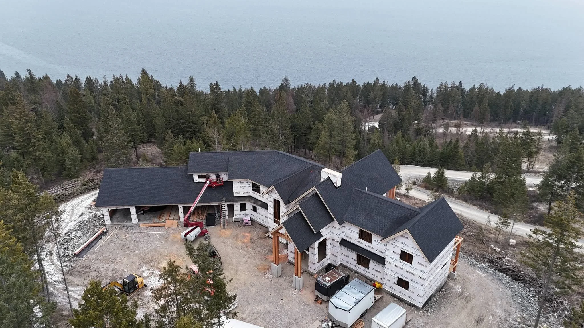 A house under construction in a forested area, with roofing being installed and construction equipment around