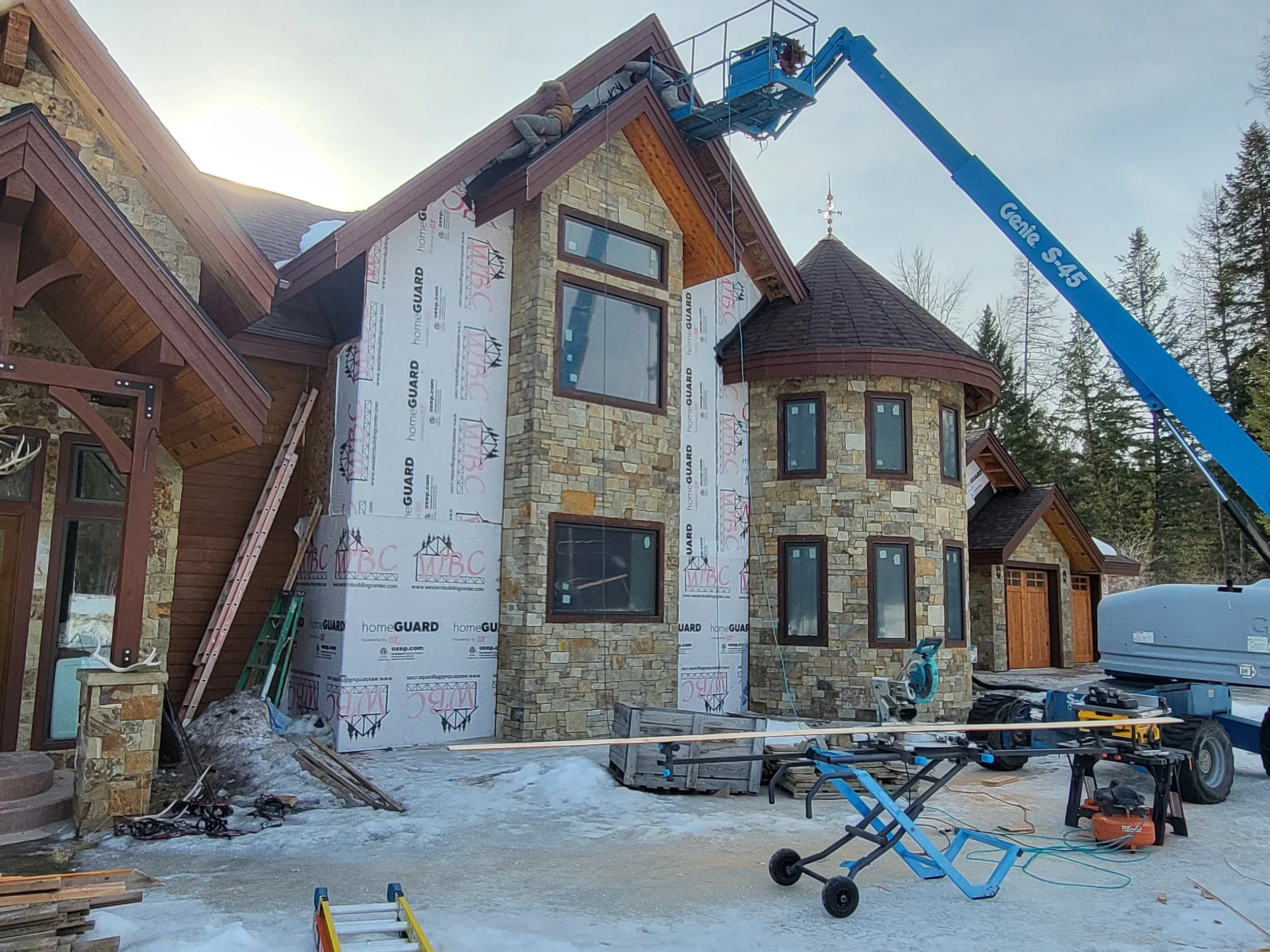 Construction workers are building a house, with one worker on an elevated lift working near the roof, and various construction tools and materials on the ground.