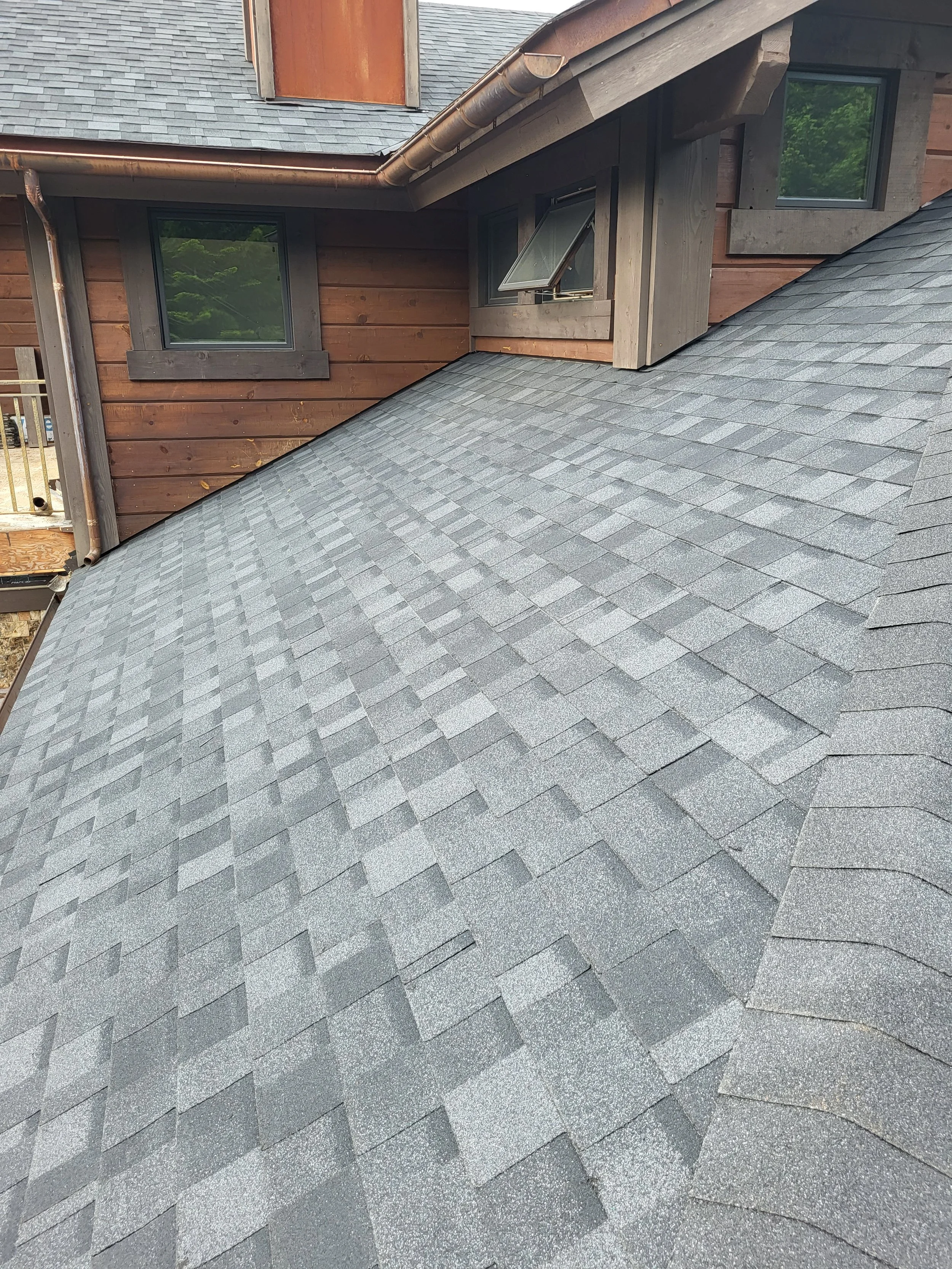 Close-up of a gray shingled roof on a house with brown wooden siding and multiple windows, including an open window.