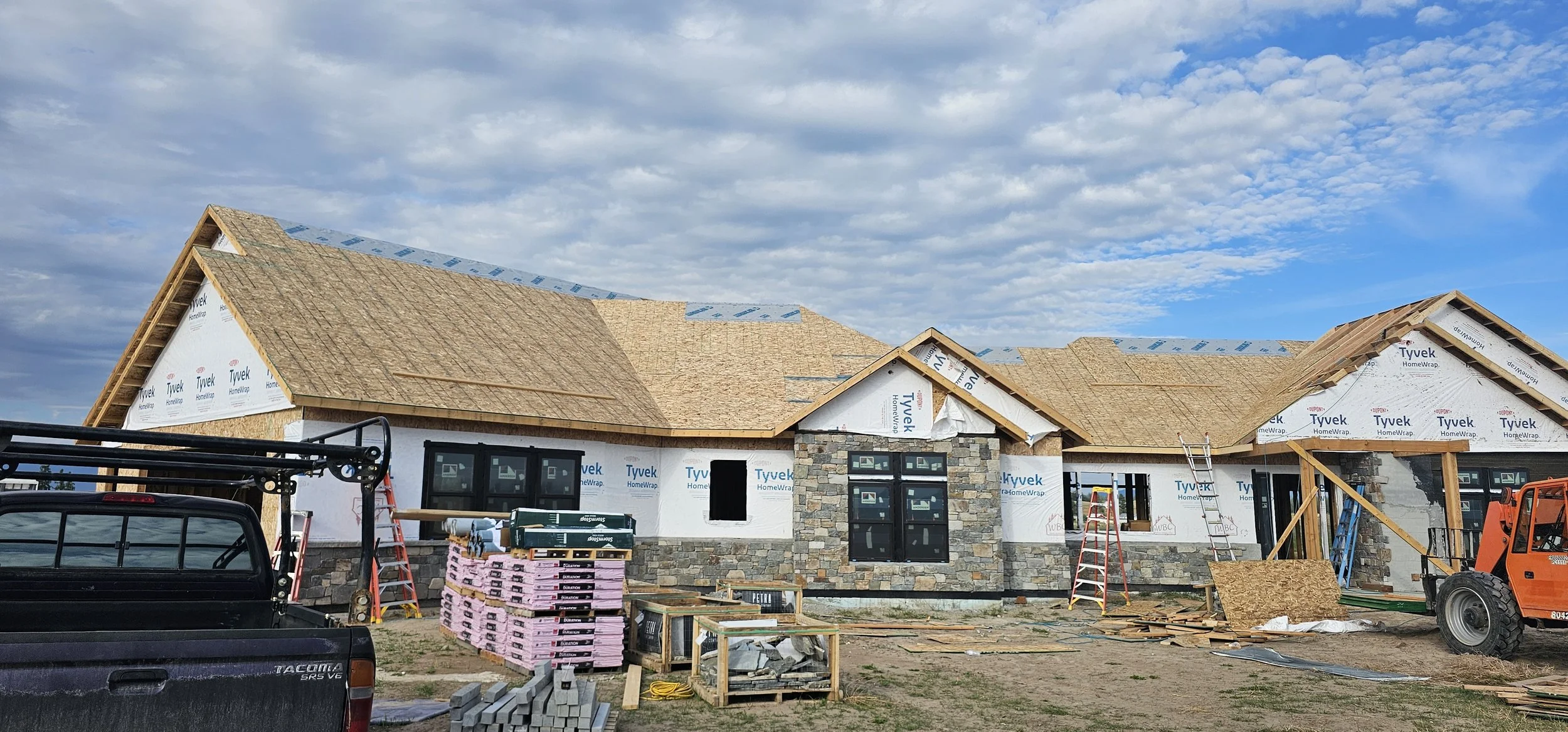 Under construction house with a partially installed roof, construction materials, ladders, and construction vehicles on site.