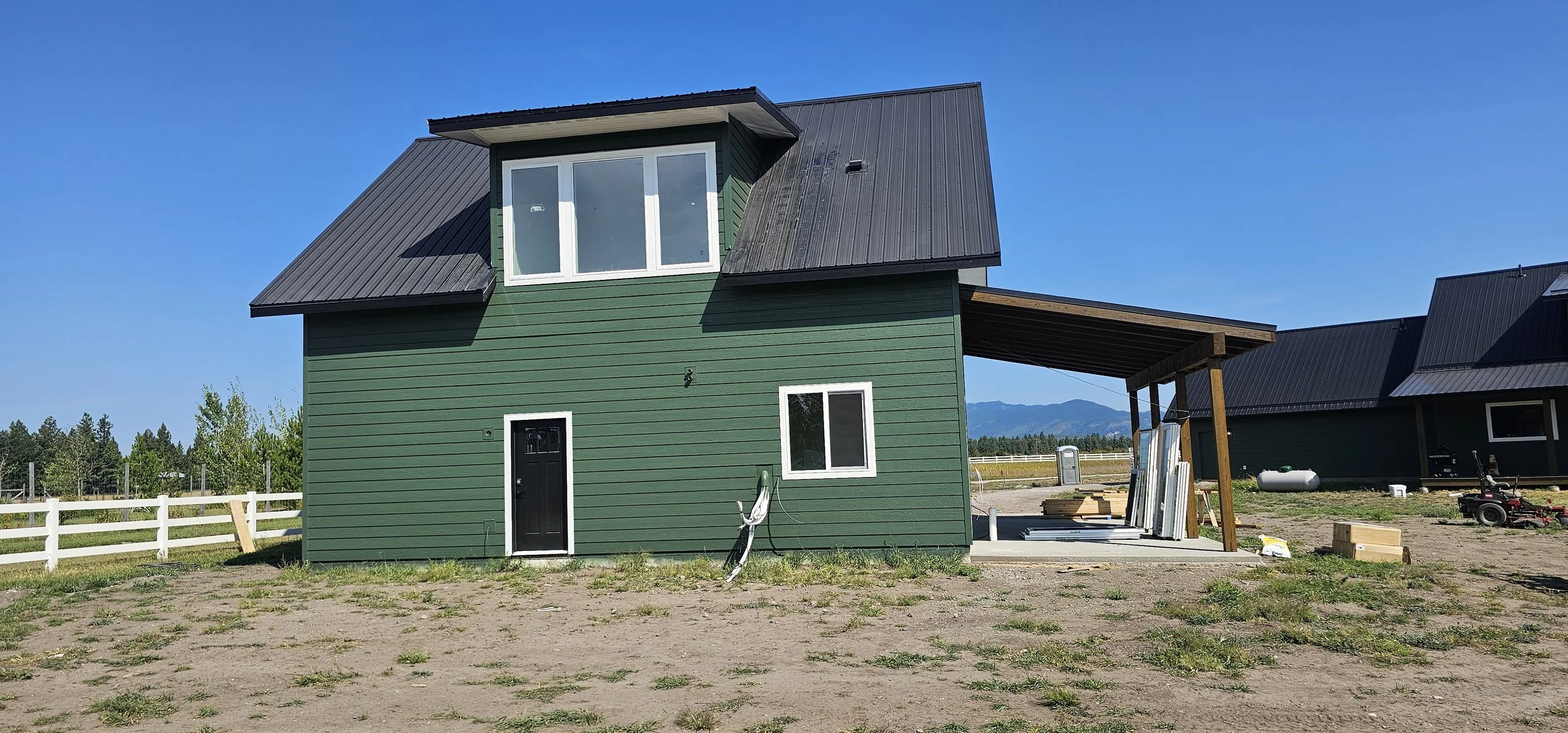 A two-story green house with a black door, white-framed windows, and a black metal roof, situated in a rural area with grass and dirt ground, and a white fence in the background.