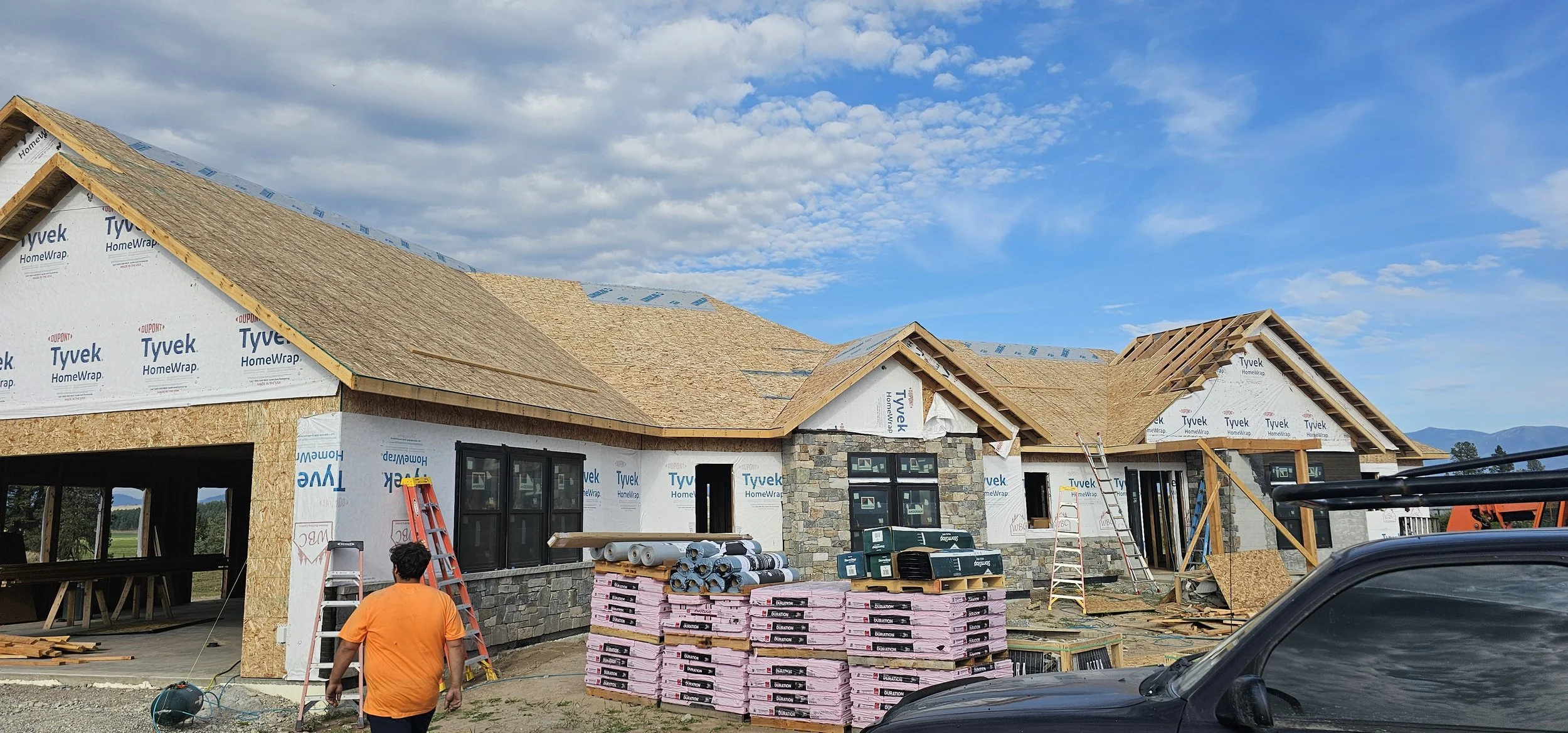 Construction site of a single-story house with a partially completed wooden roof, windows, and stone exterior walls. Building materials like insulation and plywood are stacked nearby. Construction workers are present, and ladders are in place. A blac
