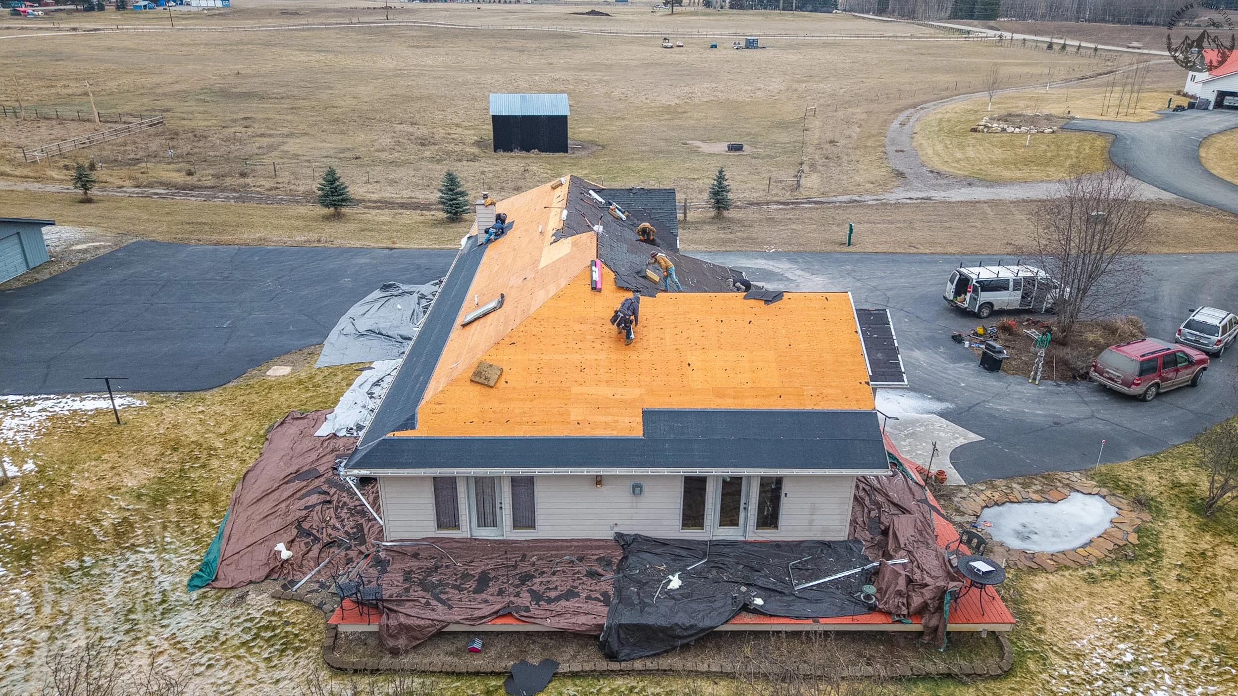 Aerial view of a house under construction with roofing workers installing a new roof, surrounded by construction materials and equipment, in a rural area with open fields and parked vehicles nearby.