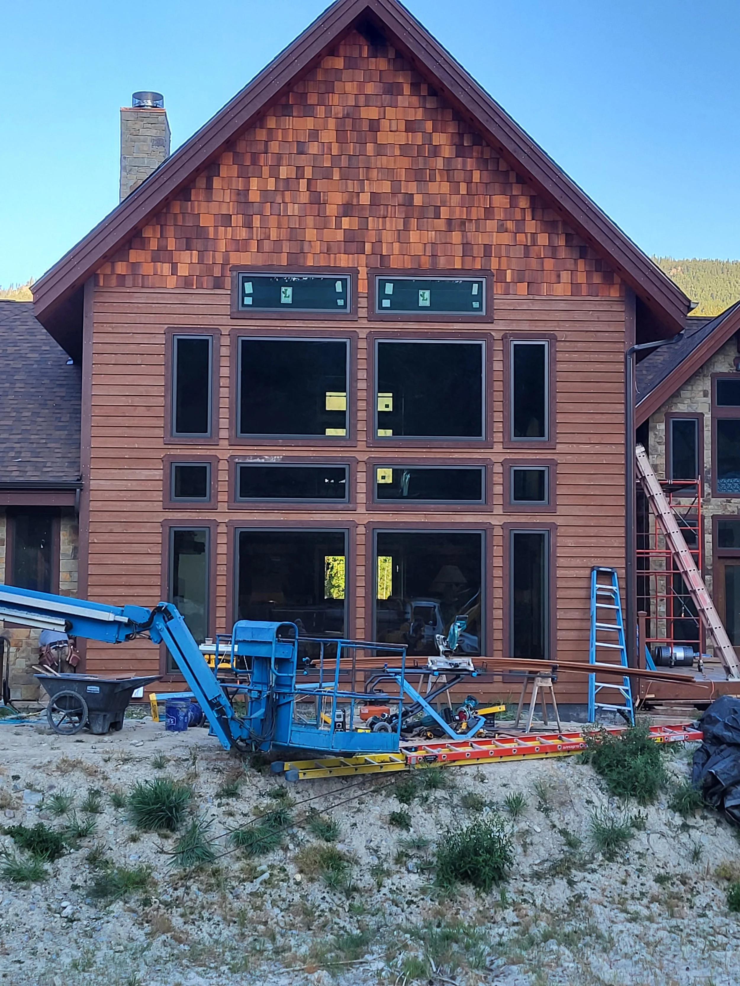 Construction site of a house with a wooden exterior, showing large windows installed on an upper and middle floor, scaffolding and construction equipment in the foreground.
