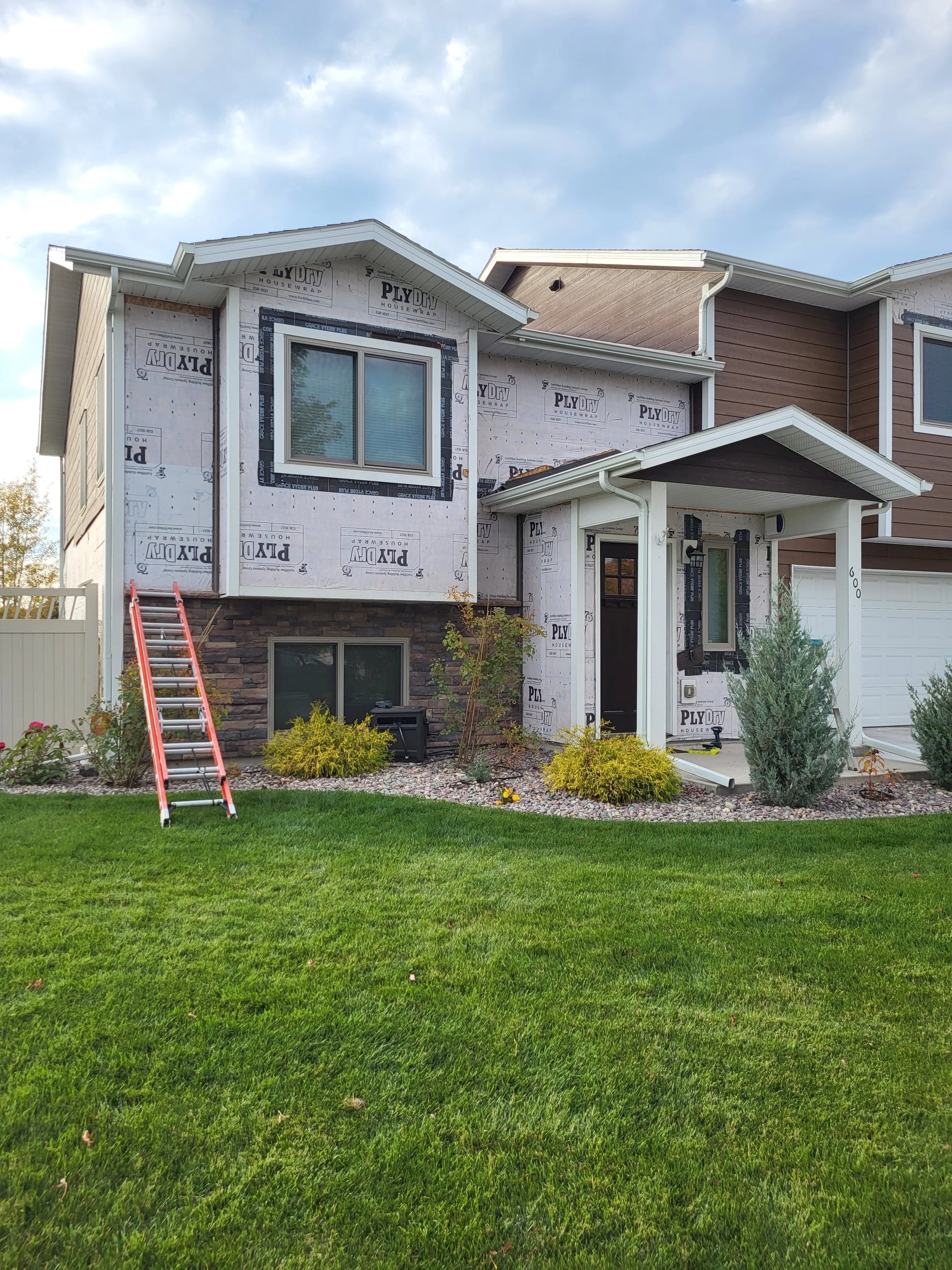 A two-story house under construction, partially covered with PlyDry house wrap, with a front yard featuring green grass, bushes, and small trees. A red ladder leans against the house near an upper window.