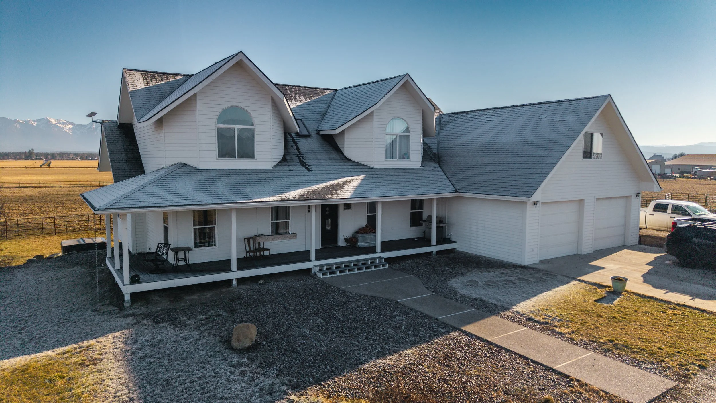 White two-story house with a large front porch, dark roof, and attached garage, set in a rural landscape with fields and mountains in the background.