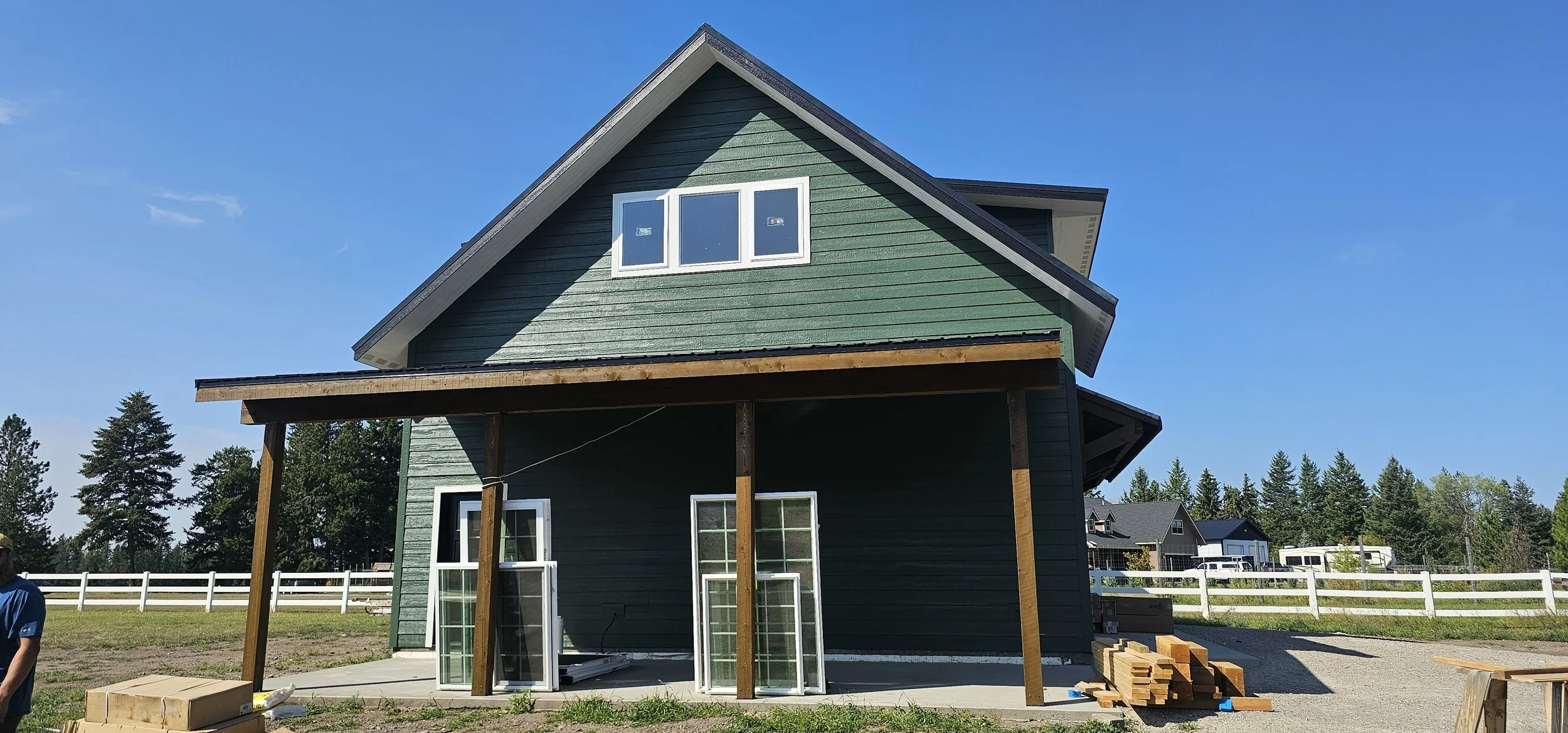 New two-story house under construction with green siding, white window frames, and a covered front porch supported by wooden beams. Construction materials and windows are on the ground nearby, and a white fence surrounds the property. Other houses an