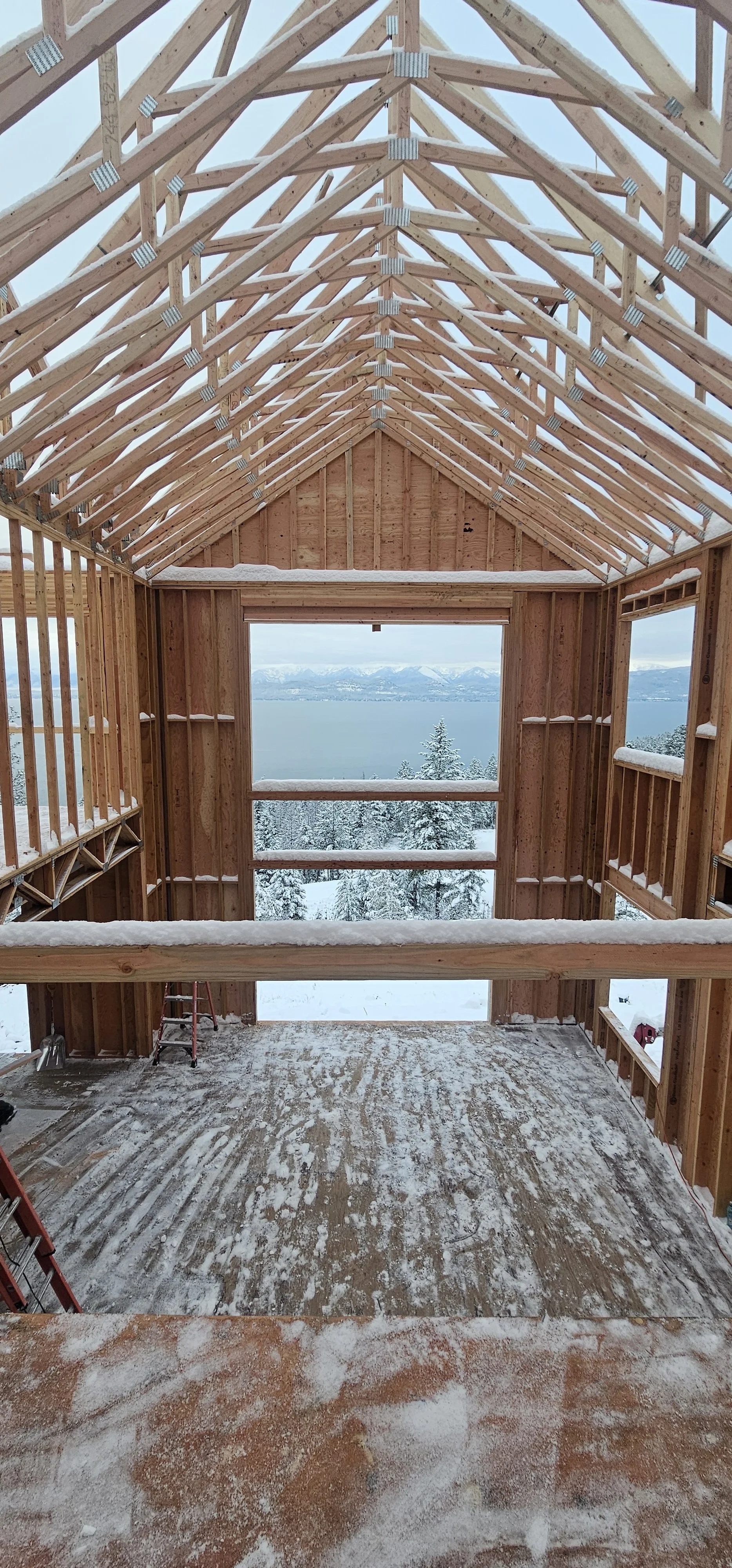 Interior view of a wooden house under construction with a view of snow-covered trees, mountains, and water in the distance.