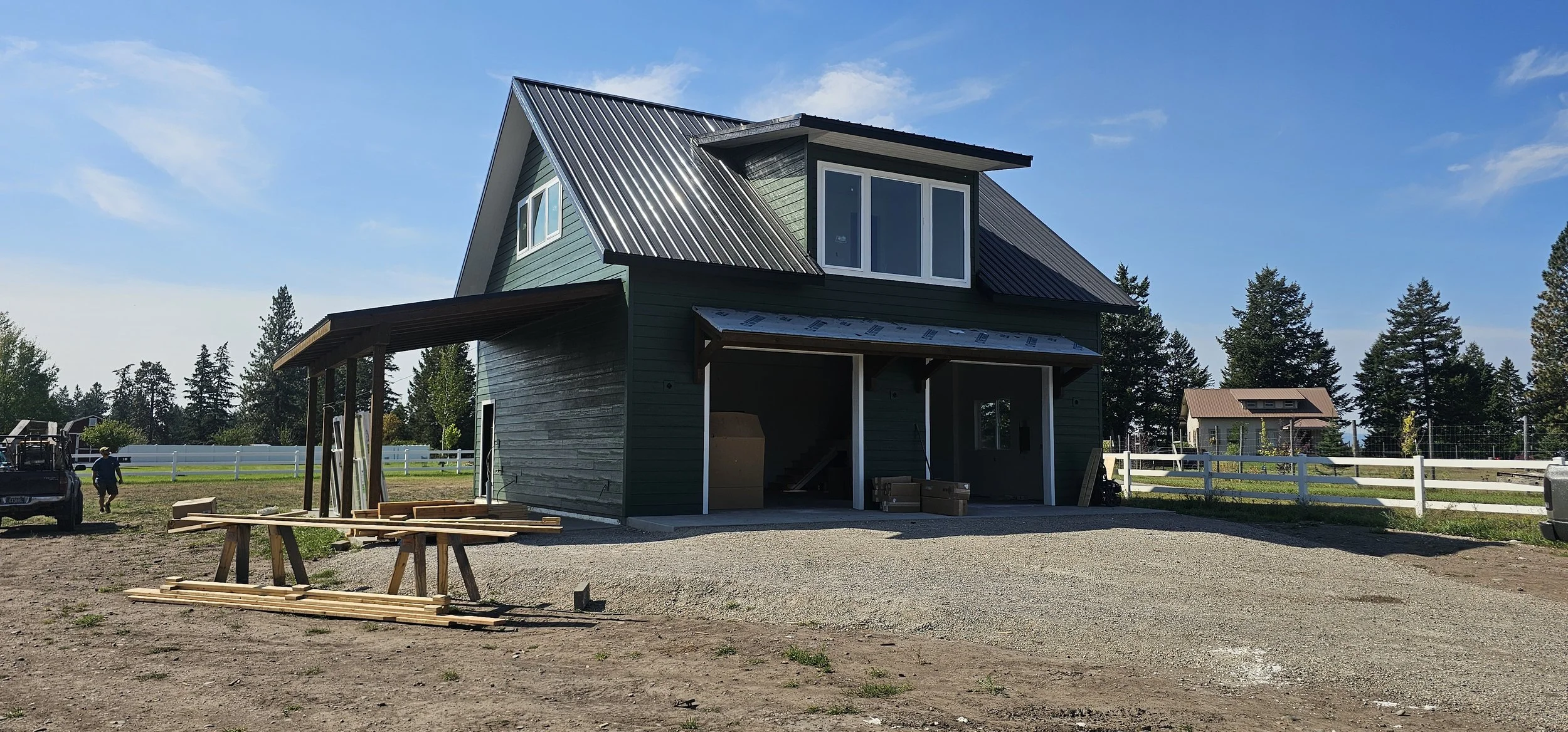 A house under construction with a green exterior, a metal roof, and large windows. Construction materials and a worker are visible in the yard, with trees and other houses in the background under a blue sky.