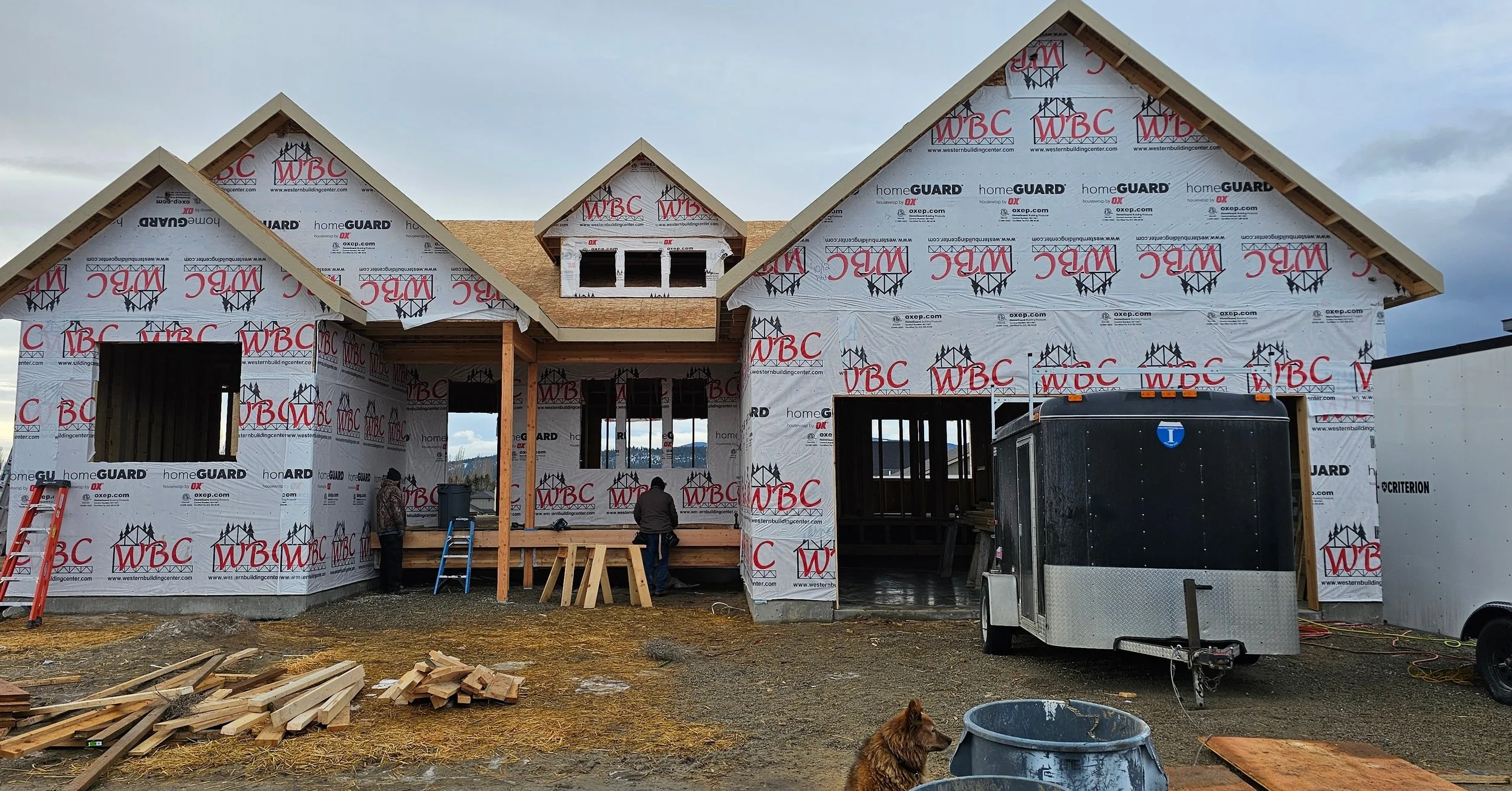 A house under construction with a wooden frame and wrapped in building material. Workers are on site, and construction equipment is visible including a trailer, a ladder, and a pile of wood. A brown dog is sitting on the ground in the foreground.
