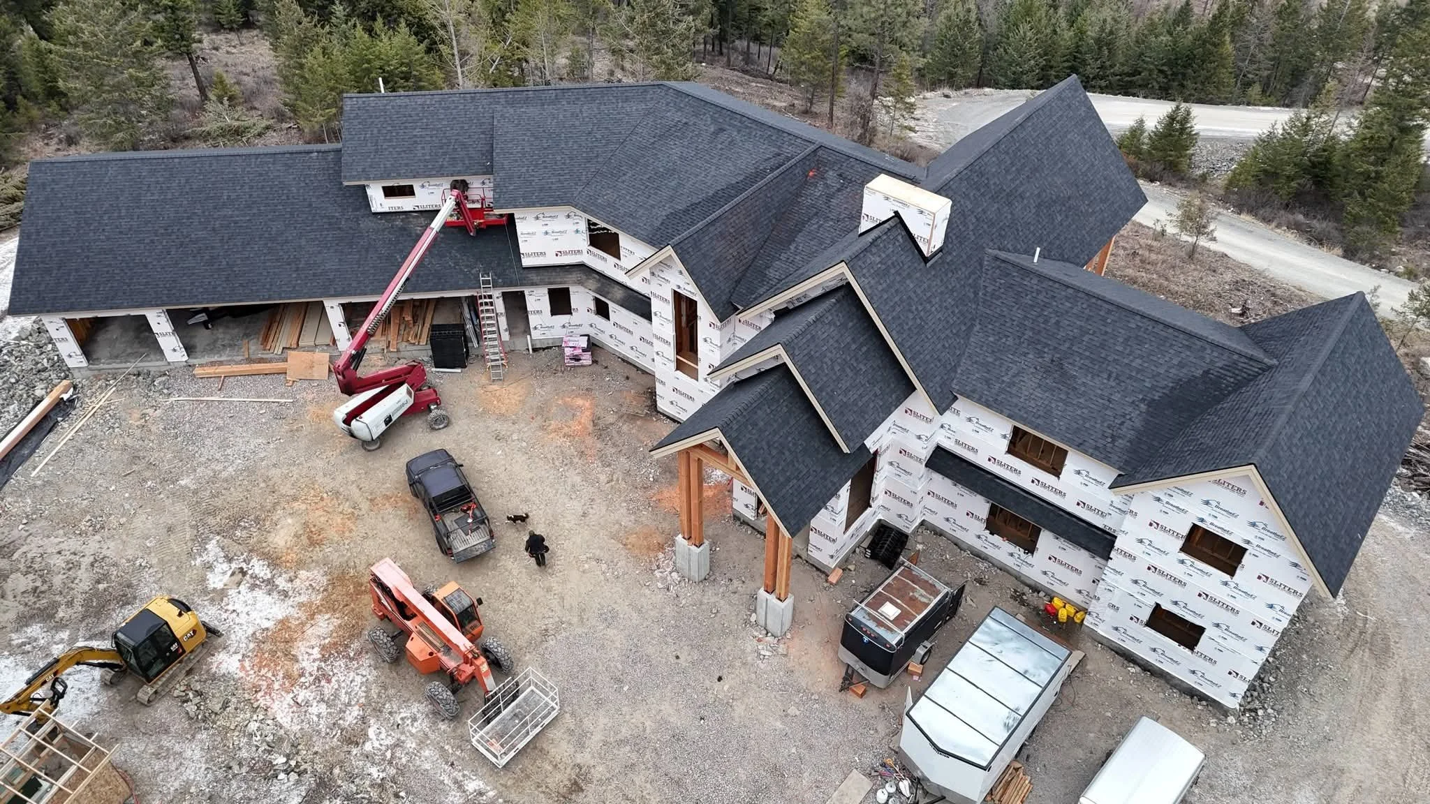 An aerial view of a house under construction with black shingle roof, scaffolding, construction equipment, and workers on site, surrounded by trees and a dirt driveway.