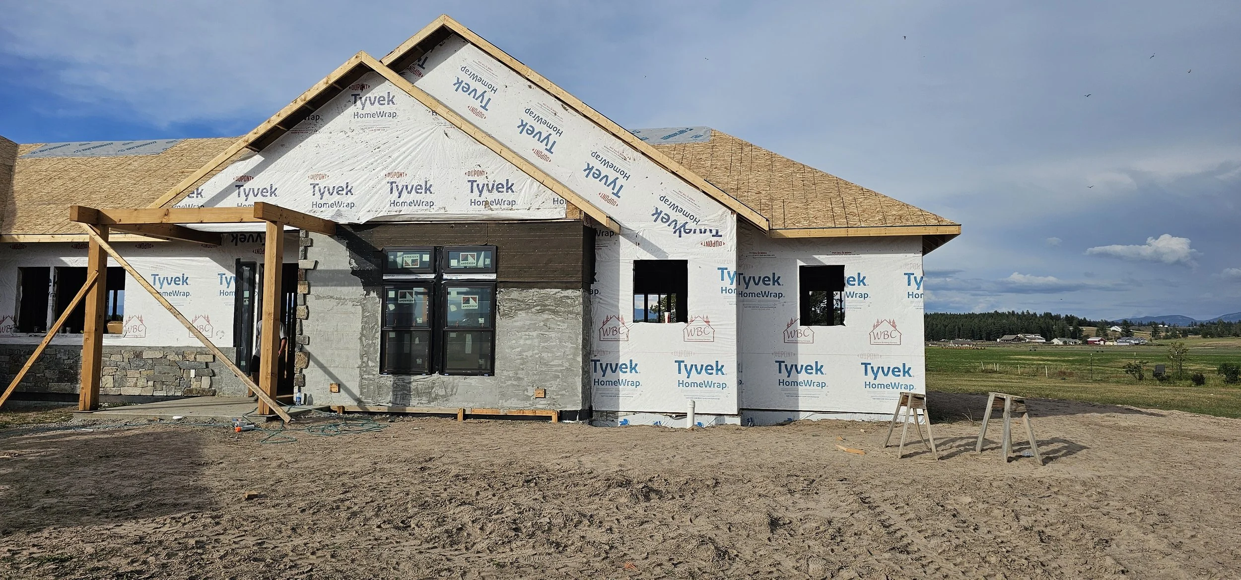 Construction site of a house with framing and exterior sheathing, windows installed, and a pitched roof in progress, with a vast open field and blue sky in the background.