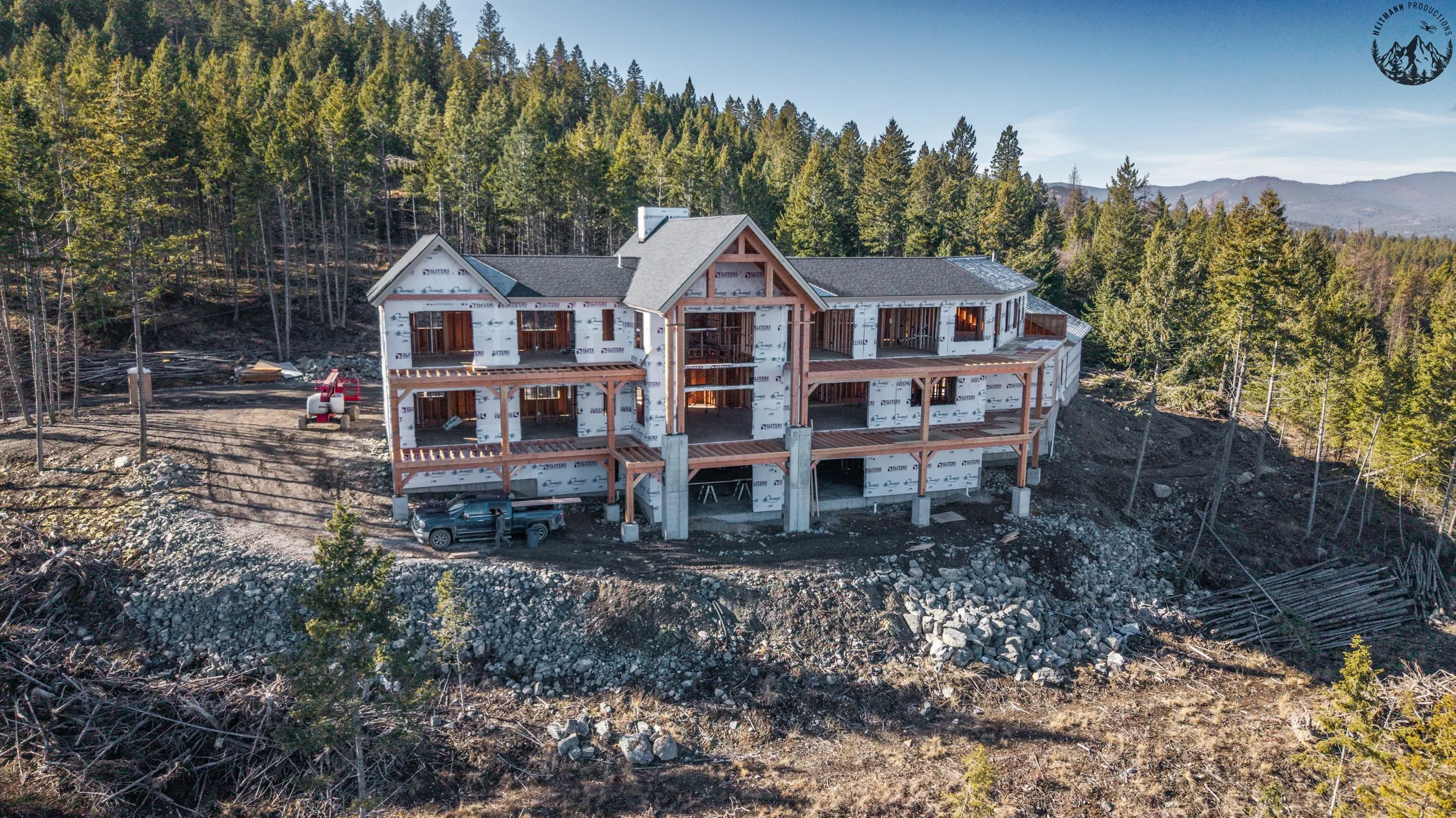 Under-construction multi-story house on a hillside surrounded by forest with mountains in the background.