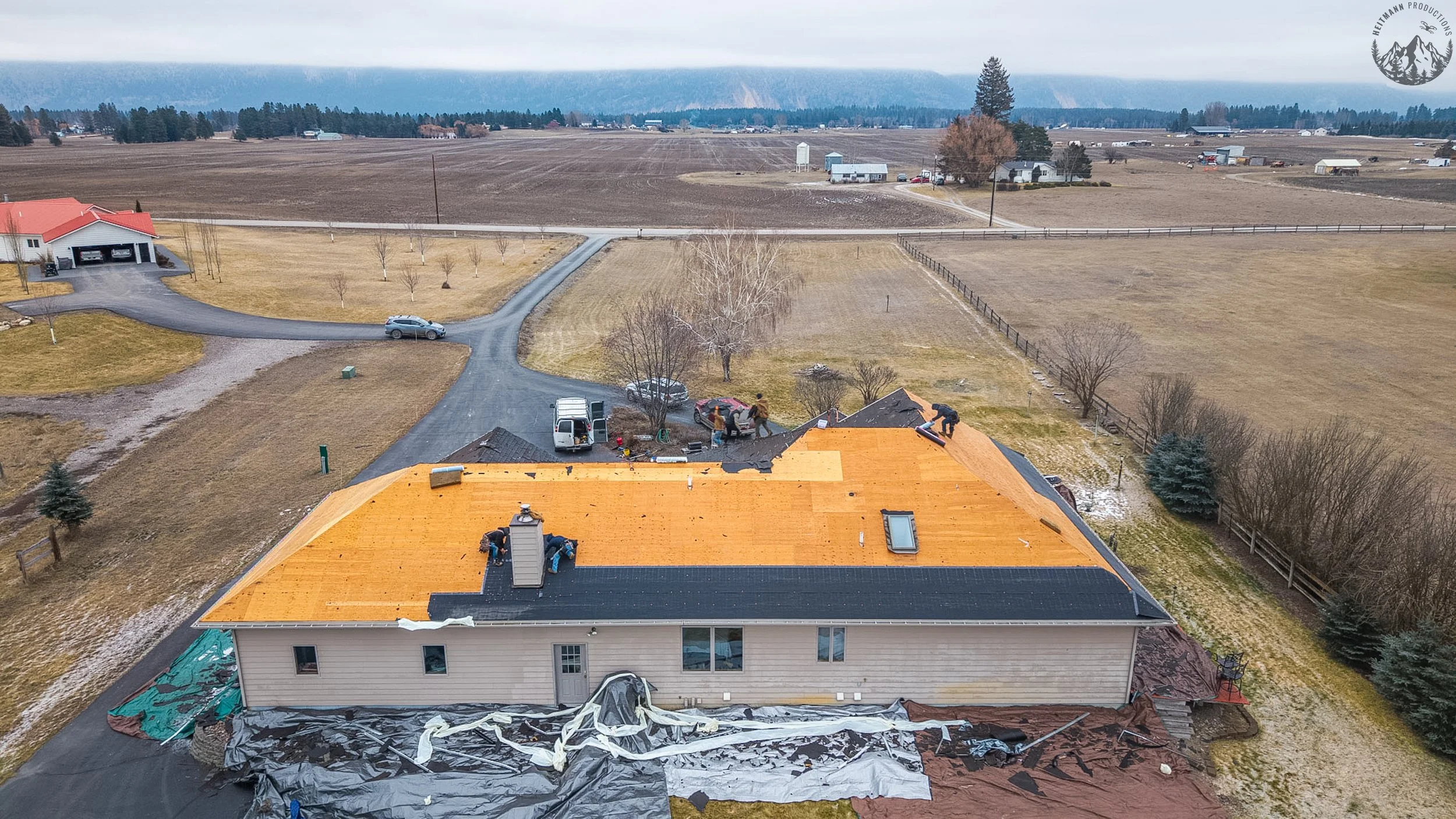 Aerial view of a house with a new orange roof under construction, workers are on the roof, with trucks and tools nearby, surrounded by a rural landscape with fields, trees, and other farm structures in the background.
