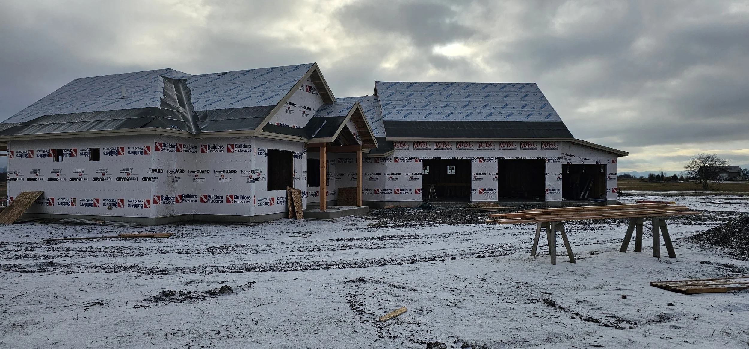 Under-construction house with snow on the ground, showing partially completed walls and roof, with construction materials and wooden beams around the site.