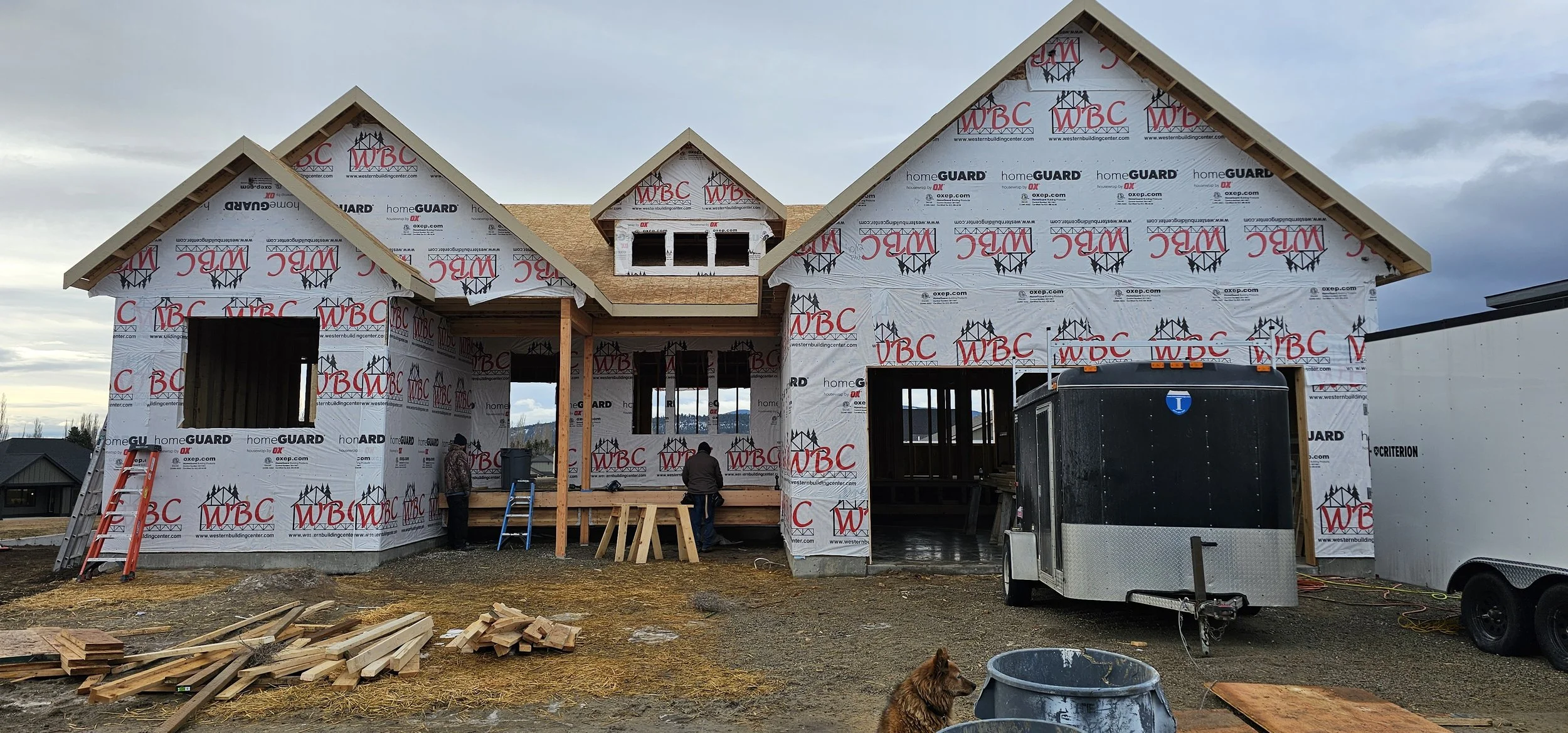 Under-construction house with framing and partial roof, construction workers, ladders, construction materials, and a dog at a building site.