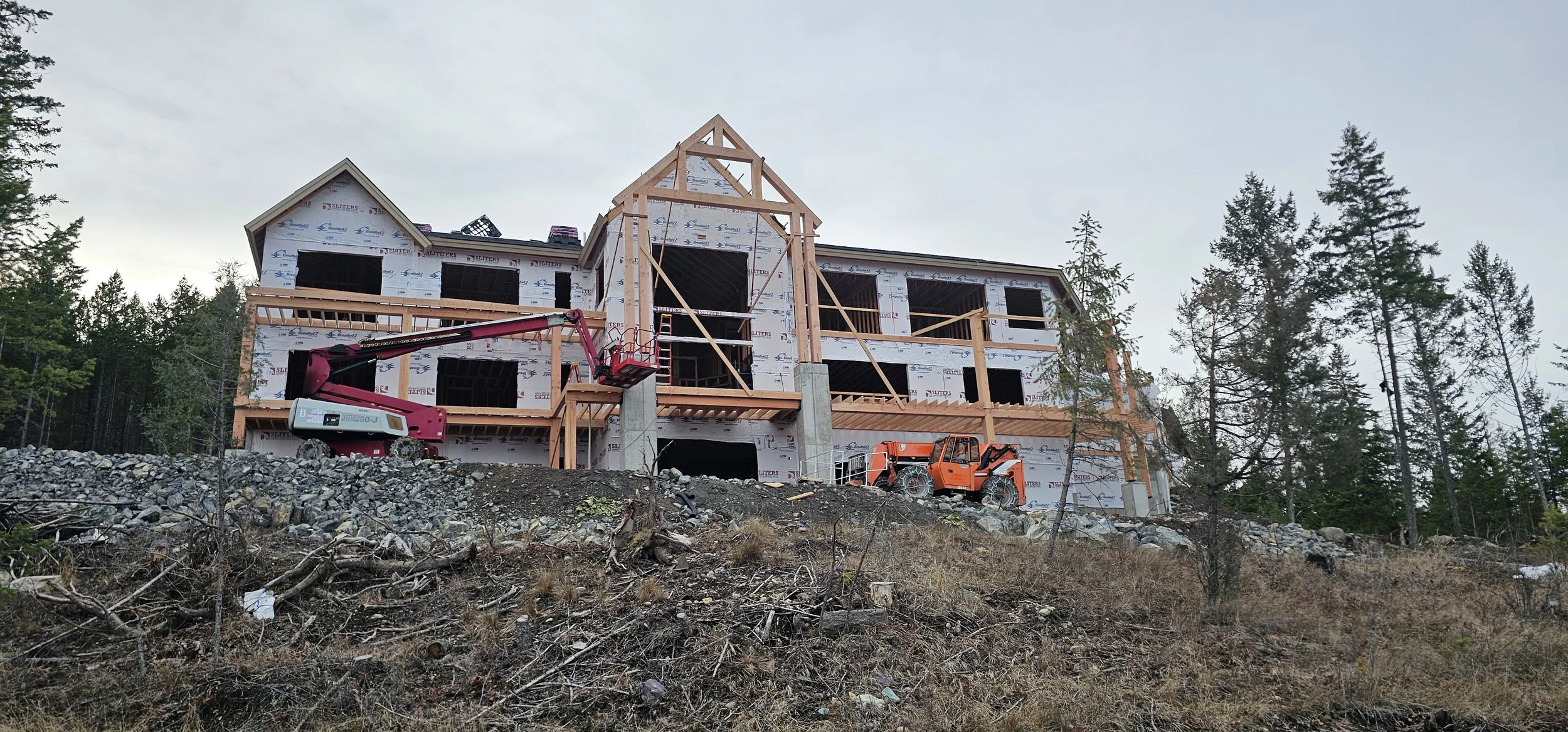 The image shows a house under construction on a rocky hillside surrounded by trees. The house frame is partly built with wooden beams and has a steeply pitched roof structure. Construction equipment, including a cherry picker and a small orange utility vehicle, is present near the building.