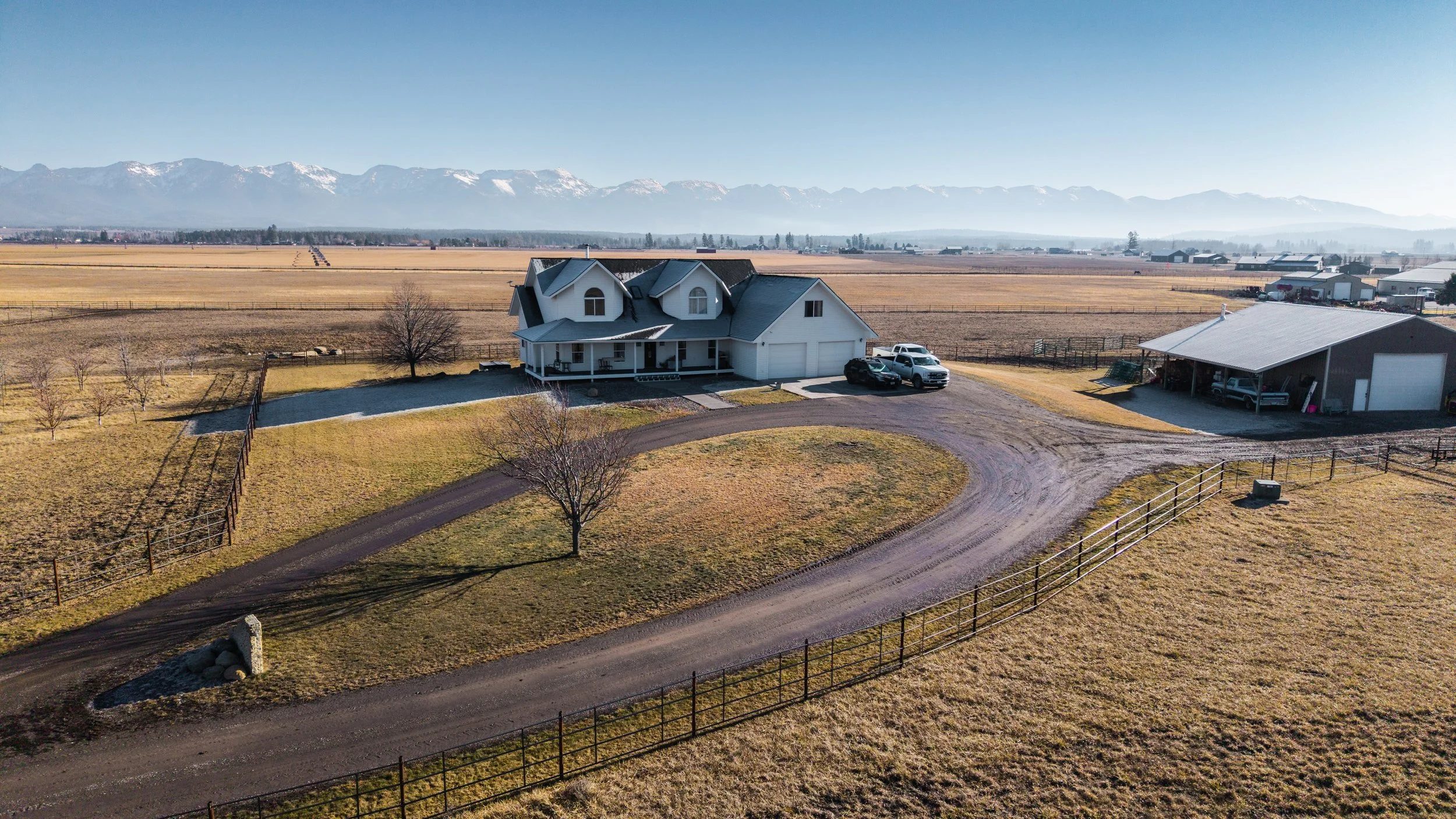 A house with a driveway leading up to it, surrounded by a fence and open fields with mountains in the background.
