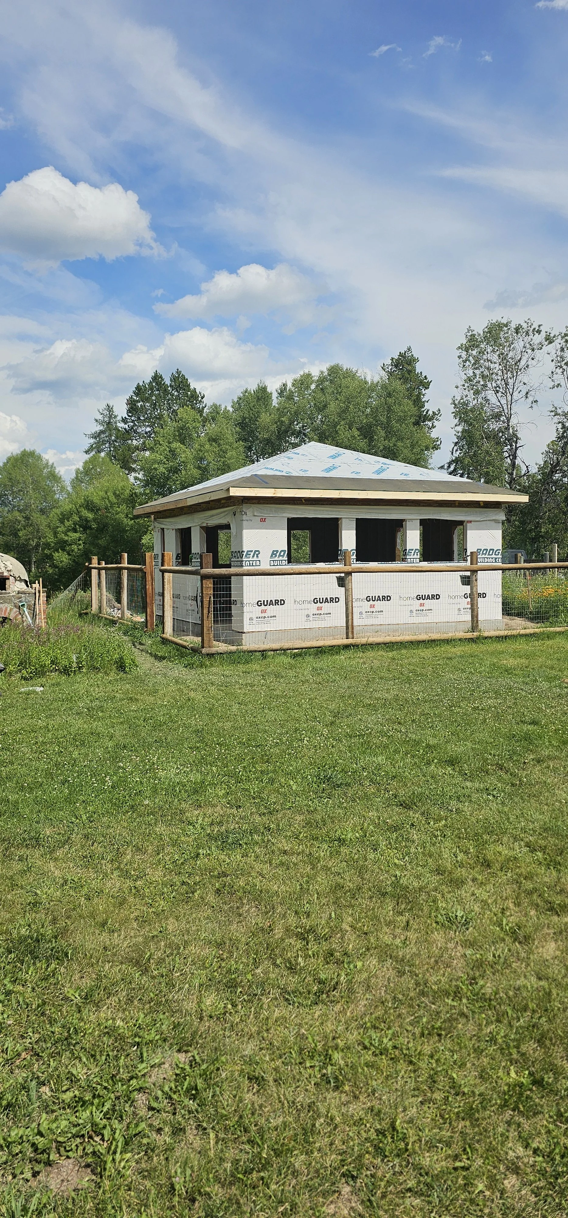 A small house under construction with a wooden fence around it, surrounded by green grass and trees under a partly cloudy sky.