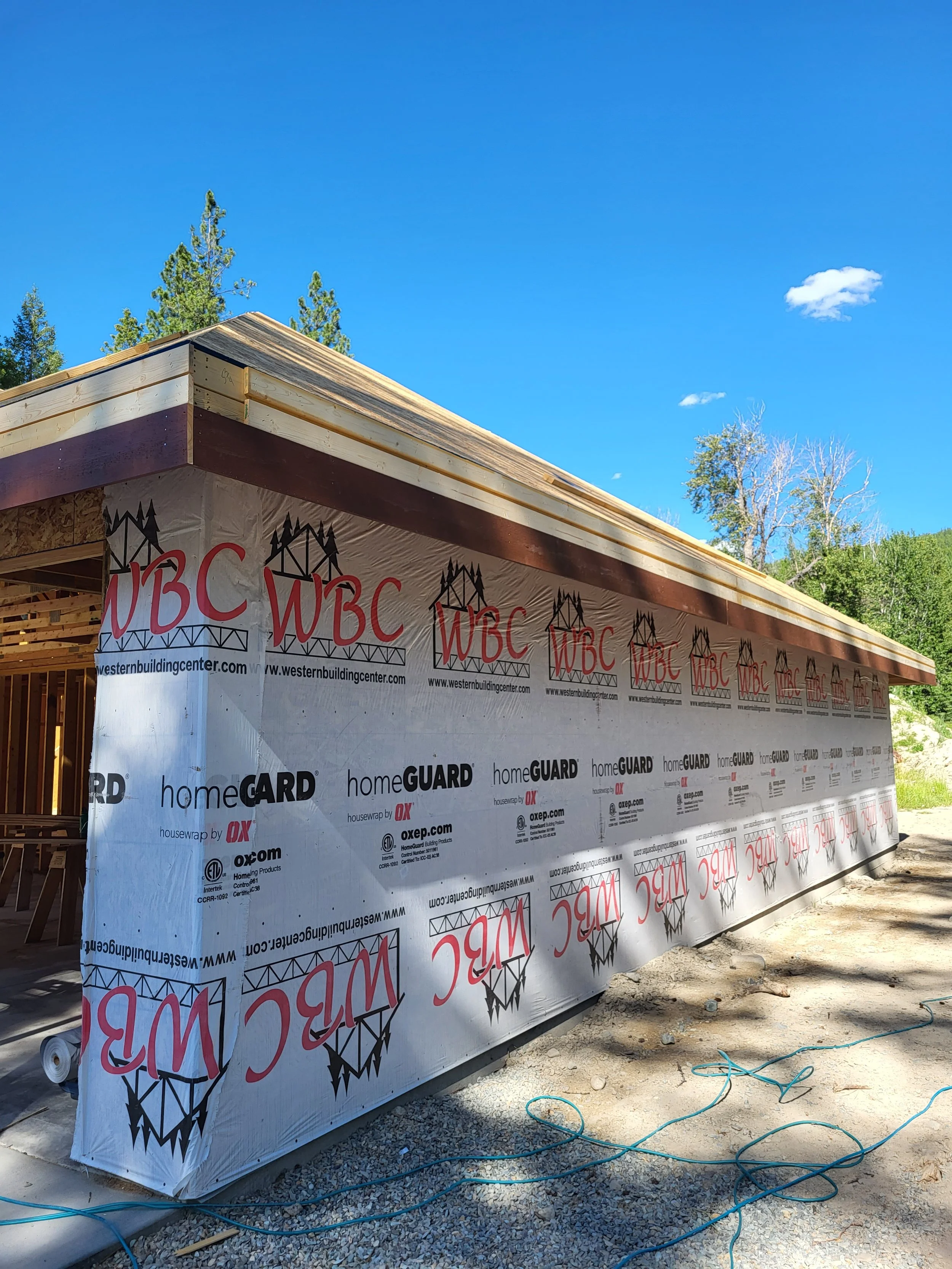 Construction site with partially built wooden house, wrapped in housewrap with logos, under a blue sky with a few clouds.