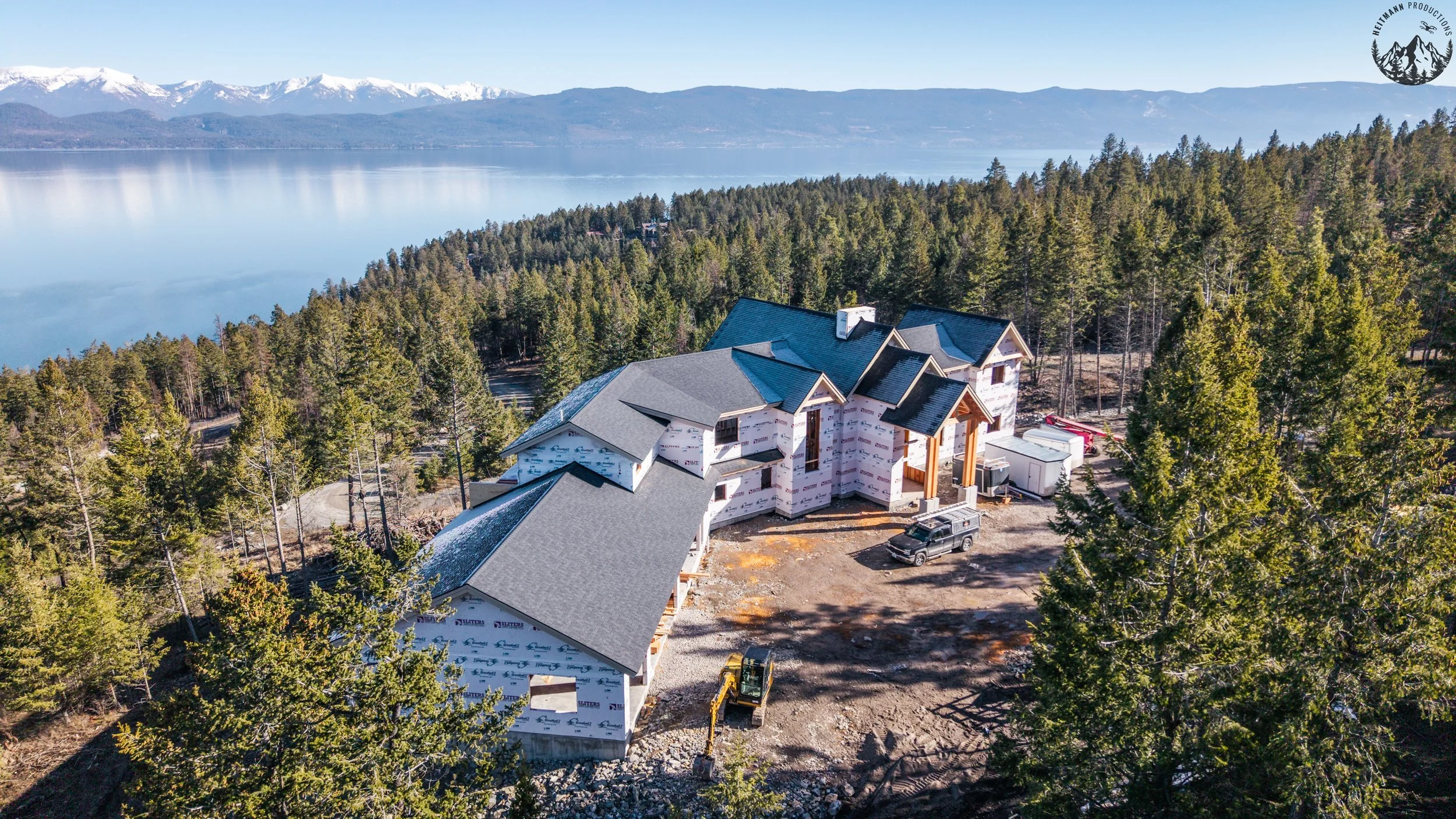 Aerial view of a house under construction surrounded by trees near a lake and mountains in the background.