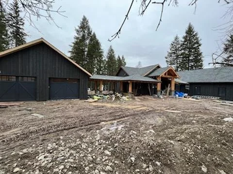 Under construction house with dark gray exterior and multiple garages, surrounded by dirt and trees in the background.